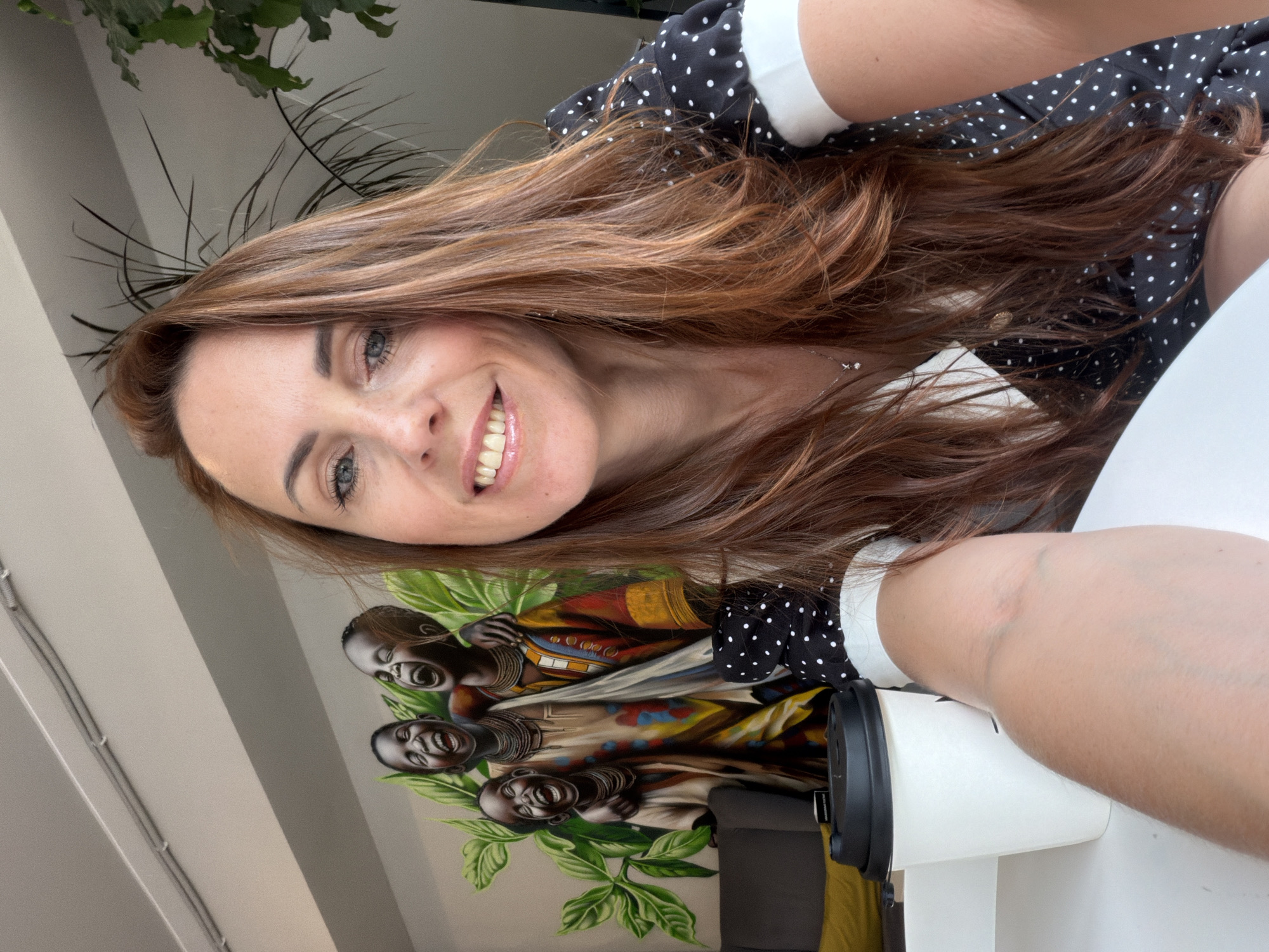 In Birobidzhan, Russia, a woman with long, reddish-brown hair is taking a selfie. She is wearing a black dress with white polka dots and a white collar, and a delicate necklace with a small pendant. Her eyes are open and she is smiling, with her teeth visible. In front of her is a white coffee cup with a black lid. Behind her is a mural depicting three children with their arms around each other, laughing and wearing colorful clothing. Lush green leaves surround the children. To the left of the mural, a dark gray couch is partially visible with a yellow pillow. A potted plant with long, thin leaves is on the right side of the frame. The environment appears to be indoors, likely a cafe or public space.