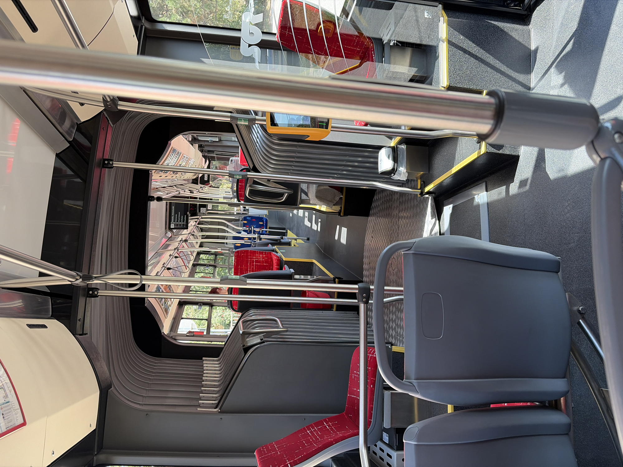 The image shows the interior of a modern public transit bus, likely a city bus, with a focus on the seating area and aisle. The seats are a mix of red with white patterned upholstery and gray plastic. Several silver metal poles for passenger support are visible throughout the aisle, extending from the floor to the ceiling. The floor is a dark gray, textured material, and there are yellow safety lines along some edges of the raised platforms.
In the foreground, two gray plastic seats are partially visible, facing forward. Further down the aisle, red seats with silver frames are prominent. One red seat on the left side is particularly clear, featuring a distinctive white crosshatch pattern. To the right of the aisle, a yellow card reader or ticket validation machine is mounted on a pole.
The back of the bus shows more red seating and the large windows that offer a view of greenery outside, suggesting an urban or suburban environment. A glimpse of a person with dark hair wearing a dark top is visible in one of the red seats further down the aisle. The architecture of the bus interior includes corrugated grey panels forming a curved section of the wall.
The image's metadata or visual cues do not provide specific information about the city of Nitra, Slovakia. However, the style of the bus, the presence of a ticket validation machine, and the general design are consistent with public transportation found in many European cities. Without further context, pinpointing the exact location within Nitra is not possible.