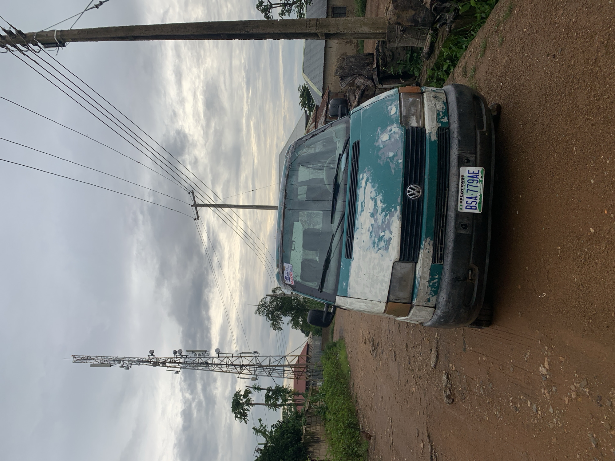 In Rafin Par, Nigeria, a light green and white Volkswagen van is parked on a dirt road. The van's paint is peeling, revealing a patch of primer underneath. The van has a license plate that reads "IIOPLATEAU BSA-779AE." In the background, a tall cell tower stands against a cloudy sky. Several power lines run across the frame, supported by wooden poles. Adjacent to the road are a few buildings with corrugated metal roofs, and lush green vegetation. A small chicken is visible on the left side of the road.