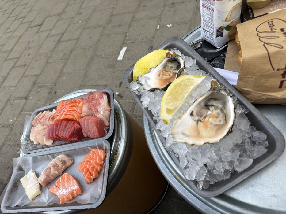 The image displays an outdoor food setup featuring raw seafood and bakery items, likely situated on a street or market pavement in Tel-Aviv, Israel. In the upper portion, two gray rectangular foam trays contain raw fish. The left tray holds larger cuts of salmon (orange-pink) and a pale white fish. The right tray presents an organized arrangement of sashimi-style slices, including dark red tuna, bright orange salmon, and light pink/white fish. Both trays rest on a round, silver-colored metallic surface, which is supported by a brown cylindrical base.
In the lower right section, a third gray foam tray features two freshly shucked oysters, each garnished with a lemon wedge, set on a bed of crushed ice. This oyster tray is positioned on a separate, lower round metallic surface. Adjacent to the oyster tray, a brown paper bag, partially visible, displays the Hebrew word "מאפיה" (Bakery) and contains baked goods. To its right, a white carton with blue labeling and Hebrew text, possibly a coconut-based product, is also partially visible. The background is composed of gray interlocking paving stones, typical of urban sidewalks or market areas. The Hebrew text on the packaging confirms the location within Tel-Aviv, Israel.