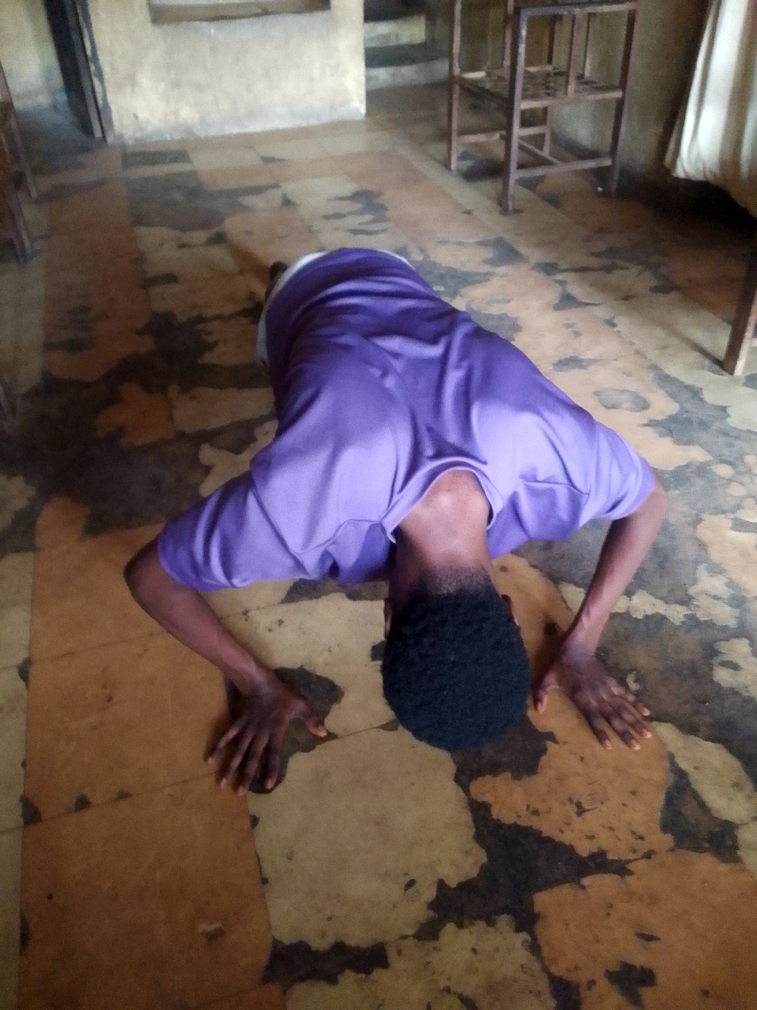An individual is positioned in a prone position on a weathered tiled floor, suggestive of performing push-ups. The person is wearing a purple shirt and appears to be in a room containing chairs and other furnishings. The exact location is unknown.