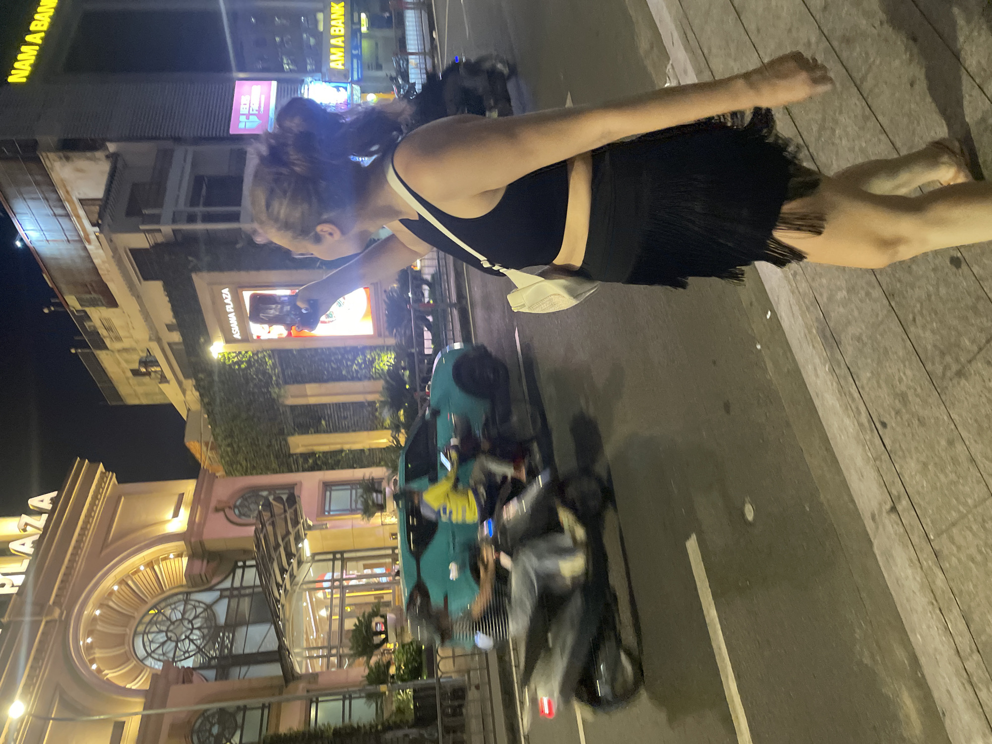 An image taken at night shows a woman standing on a sidewalk with her back to the camera, holding a smartphone to take a photo. She is wearing a black tank top and a short, black fringe skirt. She has her hair tied up in a ponytail and is carrying a white purse over her shoulder. In the background, there are illuminated buildings and streetlights. One building displays a sign that reads "NAM A BANK." Another sign reads "ASIANA PLAZA." Vehicles, including motorcycles and a teal car, are moving on the street, indicating it is a busy urban area. The scene is likely in Thủ Đức City, Vietnam, given the visible signs and urban setting.