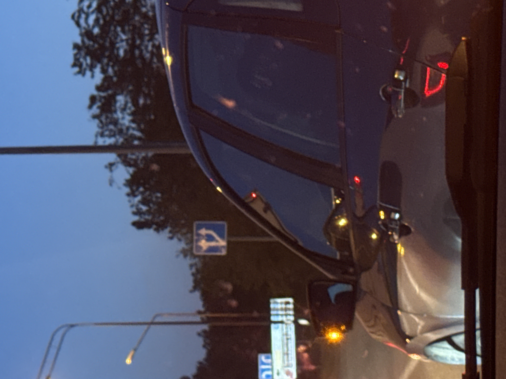 The image shows the view from inside a car, with the focus on another car parked beside it. The second car is dark blue with a silver reflection. The rear light is visible. Outside, there is a blue sign indicating a turn, and street lights are visible. The setting is likely a city street at dusk.