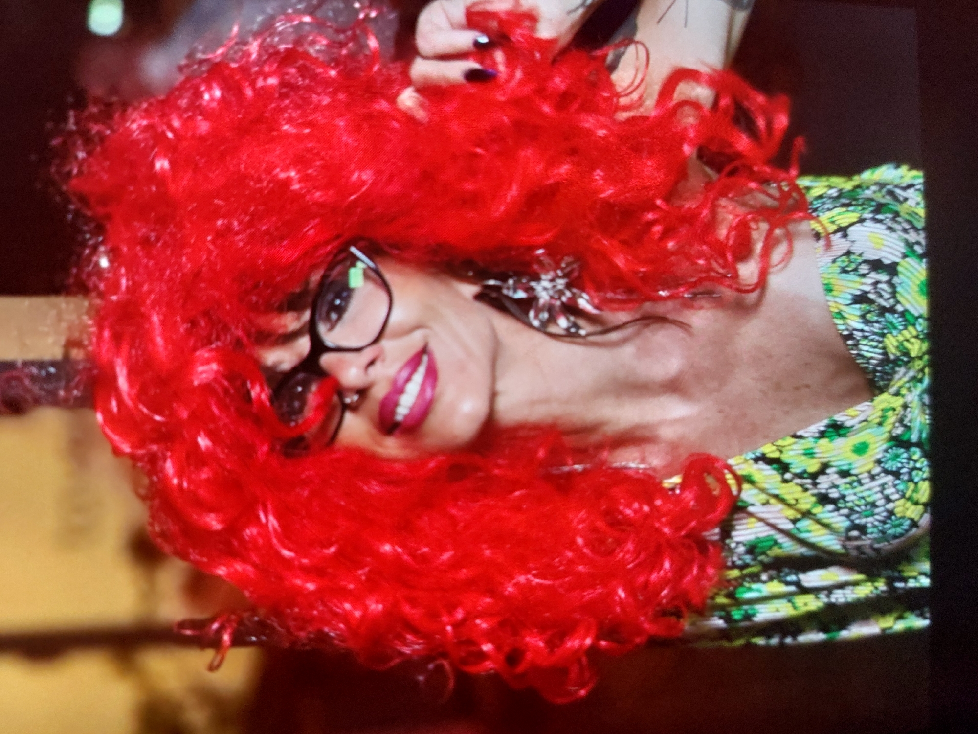 A close-up, horizontally oriented portrait features a person with striking, voluminous bright red curly hair, likely a wig. The subject is wearing black-rimmed glasses and vibrant red lipstick, smiling broadly to reveal white teeth. Her expression appears confident and engaging. A distinct, ornate metallic earring, possibly shaped like a flower or snowflake, is visible on her right ear. A hand with dark nail polish is partially visible in the upper right, seemingly interacting with her hair. The person wears a top or dress with a lively green, yellow, and white floral pattern. The background is dark and out of focus, emphasizing the subject. The setting is likely indoors, and while the location context provided is Tsarevo, Bulgaria, no specific visual cues within the image confirm this. No text is visible in the media file.