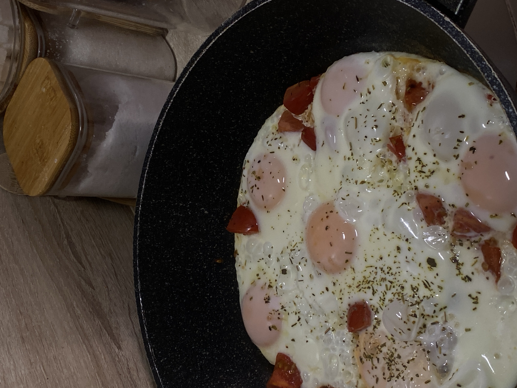 The image captures a close-up view of food cooking in a black frying pan with a speckled non-stick surface. Inside the pan, several fried eggs are visible, with set white parts and translucent, pale orange yolks. Diced red tomatoes are distributed throughout the eggs. A substantial amount of dried herbs, resembling oregano or a mixed spice, is sprinkled over the entire dish.
The pan rests on a light-colored wooden surface, likely a countertop or table, which exhibits a subtle grain pattern. In the upper left background, two clear glass jars with light wooden lids are partially visible. One jar contains a white granular substance, possibly salt or sugar.
The scene depicts a domestic cooking activity, with the eggs appearing mostly cooked. The lighting suggests an indoor setting. No people are visible, and there is no discernible text within the image. The overall impression is of a simple, homemade meal being prepared.