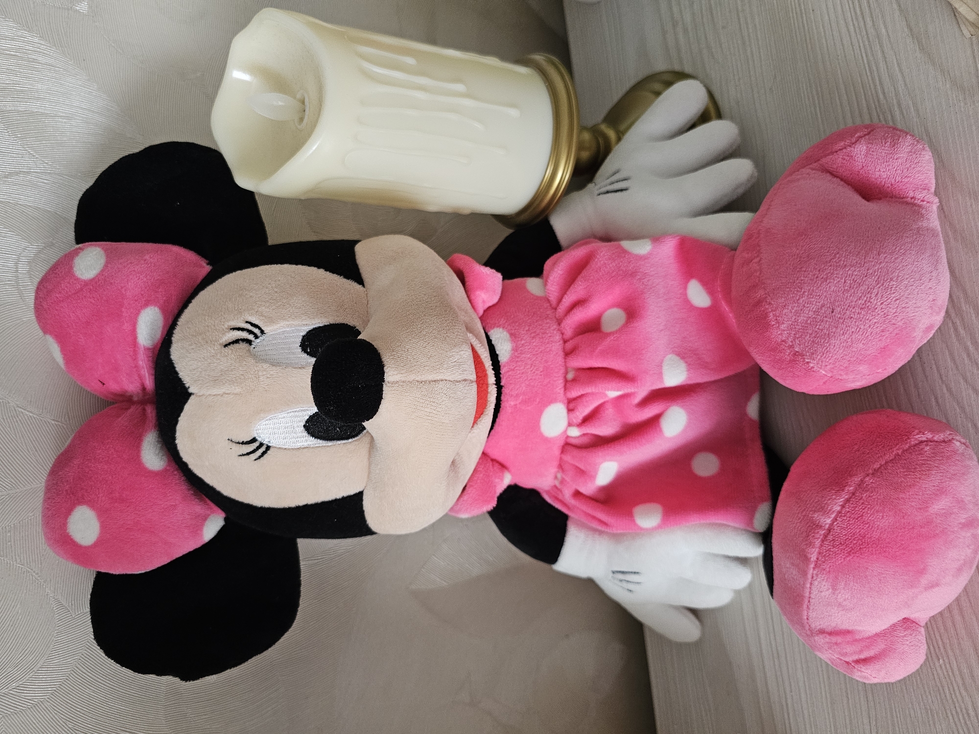 A plush Minnie Mouse toy lies on a textured surface, possibly a bedspread or couch. The toy is primarily pink and black, with white polka dots on its ears and dress. Its eyes are closed, giving it a serene or sleepy appearance. To the left of the toy, an artificial candle with a gold base is placed on its side. The candle is a creamy white color and appears to be made of wax. The background is a light, neutral color with a subtle pattern. The scene is brightly lit, suggesting daytime indoors. There is no visible text in the image.