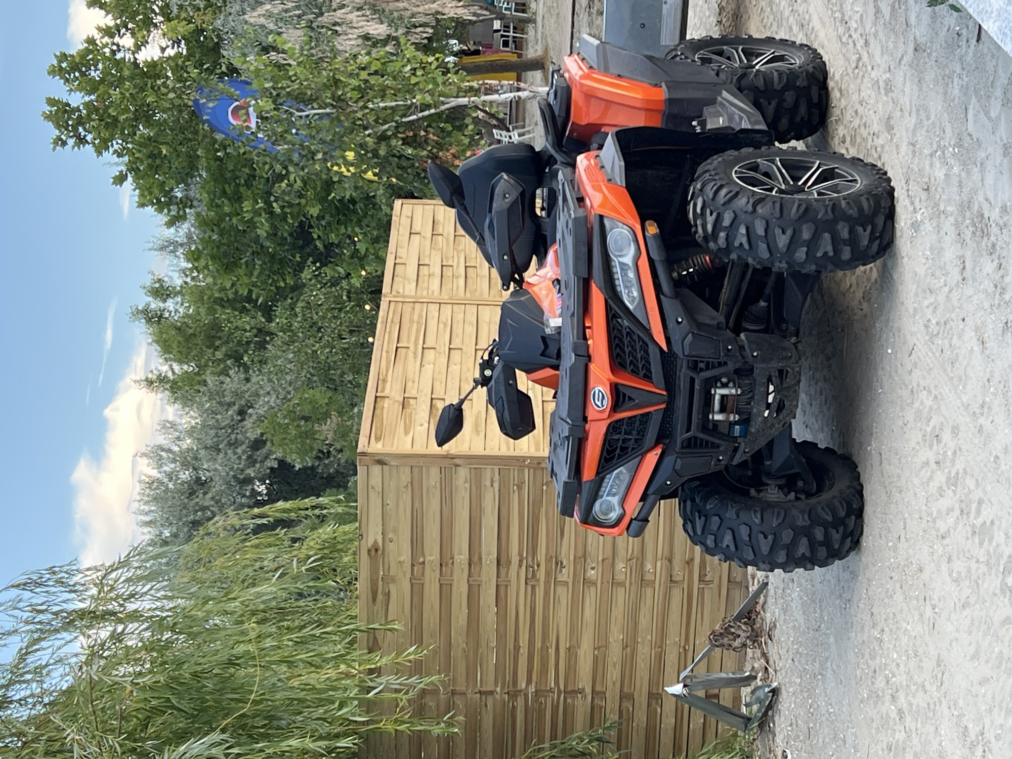 An orange and black ATV is parked on a sandy surface in front of a wooden fence. The ATV is facing forward and to the left. It has large, knobby tires, a black seat, and black handlebars. To the left of the ATV, there is a partially visible metal object with a chain. Behind the ATV is a light brown wooden fence made of horizontal planks. Above the fence, there are green trees and a blue sky with white clouds. A large weeping willow tree with long, drooping branches is on the left side of the image, in front of the fence. In the background, beyond the trees, some beach chairs and tables are visible, suggesting a coastal or resort area. The scene appears to be located in Năvodari, Romania.