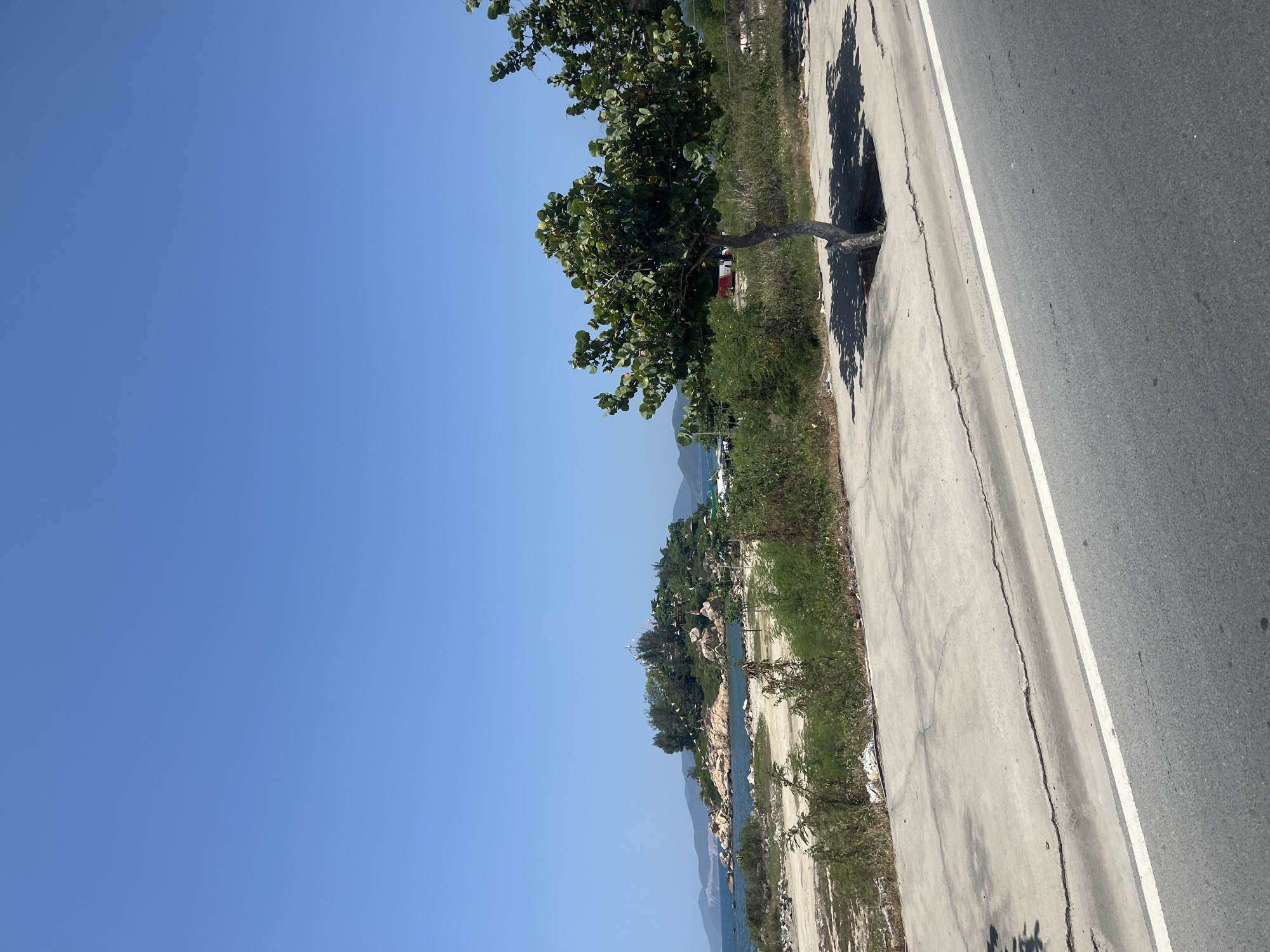The image depicts a coastal landscape with a clear blue sky. In the foreground, a cracked asphalt road with a white line runs diagonally. Adjacent to the road is a concrete walkway, also showing signs of disrepair with visible cracks. A small tree with green foliage is planted in a square cutout in the concrete walkway, casting a shadow. Beyond the walkway, there is an area of dry, sandy ground with sparse green vegetation.
In the middle ground, a body of blue water is visible, meeting a rocky coastline. A small, densely vegetated island with trees and several structures on its summit is situated in the water. The island is adorned with numerous small white flags. The background shows distant hills or mountains under the expansive sky.
The scene appears to be in a coastal area of Nha Trang, Vietnam, likely on a sunny day. There are no people or vehicles clearly visible in the image. The environment suggests a somewhat undeveloped or developing area near the coast.