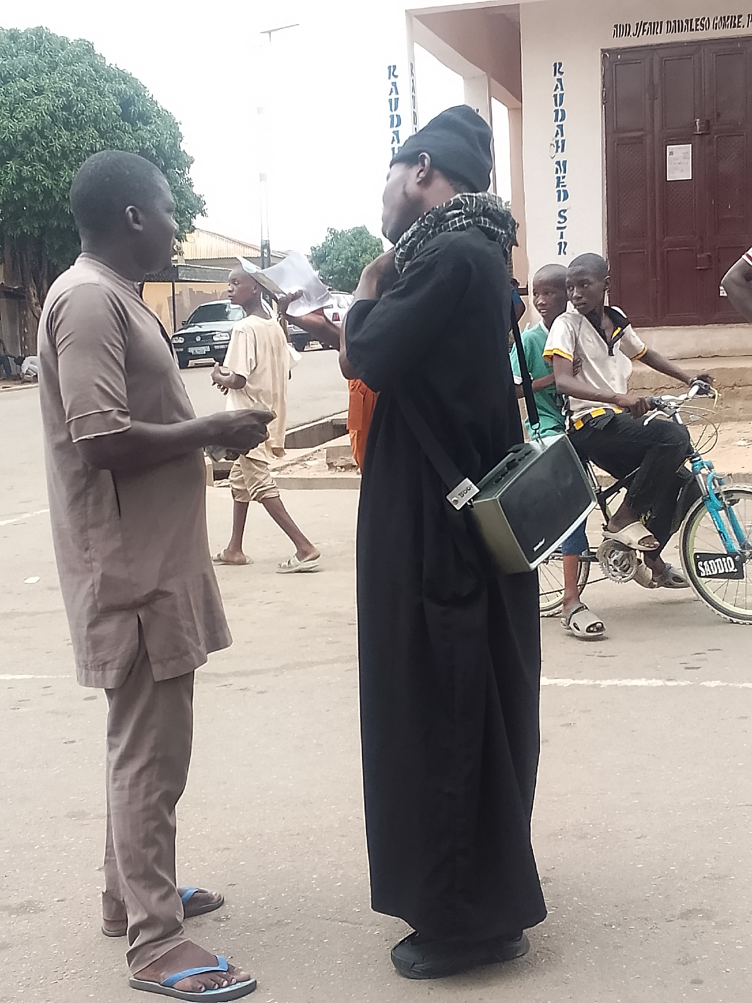 The image captures a street scene in Gombe, Nigeria. Two men are engaged in conversation in the foreground. The man on the left wears a brown, short-sleeved tunic and pants with flip-flops. The man on the right is dressed in a long black robe and cap, with a checkered scarf around his neck, and carries a rectangular bag on his shoulder. In the background, a boy sits on a bicycle, and another boy stands nearby. There is a building behind them with text "RAUDAH MED STR" visible on the wall. A car is parked on the street in the distance.