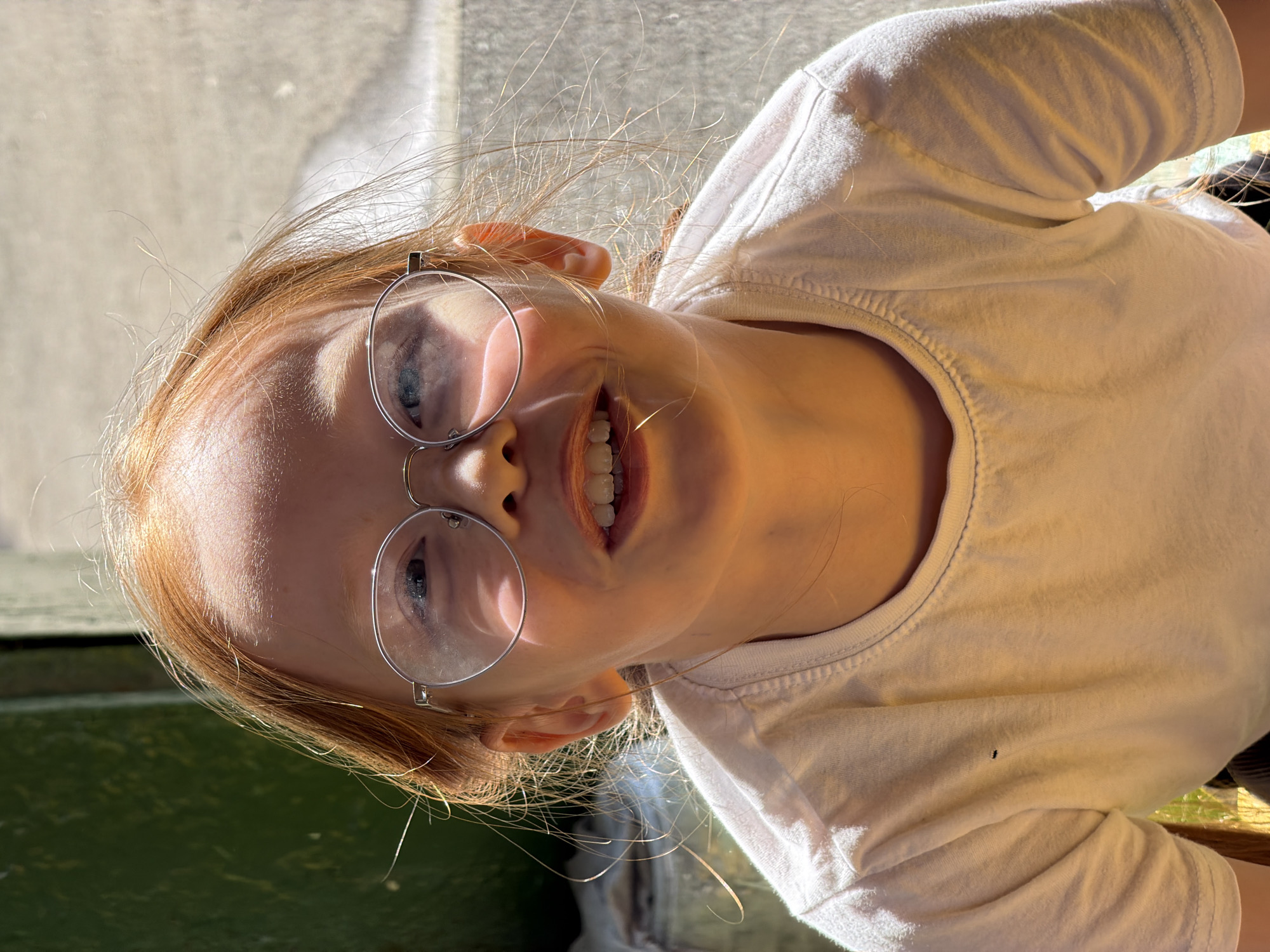 A close-up portrait shows a young child with fair skin, light red or strawberry blonde hair, and blue eyes, smiling brightly at the camera. The image is rotated 90 degrees clockwise, giving it an unconventional, playful perspective. The child is wearing round, wire-rimmed glasses, through which their eyes are clearly visible, reflecting the bright light. Their mouth is open in a wide smile, revealing white teeth.
The child is dressed in a plain white, short-sleeved t-shirt. A notable detail on the shirt is a small, dark, rectangular mark located on the front, slightly to the viewer's right. Strands of their fine hair are illuminated by strong sunlight, suggesting an outdoor setting or proximity to a window.
The background is softly blurred but distinguishable. Above the child's head, a light-colored, textured surface is visible, possibly a wall or paving. Below their chin, a dark green, somewhat textured surface appears, which could be a bench, a railing, or another painted outdoor object. The intense sunlight strongly illuminates the child's face and hair, creating clear highlights and shadows, indicating a bright, sunny daytime environment. The overall mood conveyed by the child's expression is one of happiness and engagement. No text is visible in the image. The specific location of Izhevsk, Russia, is provided as context but not visually discernible from the image itself.