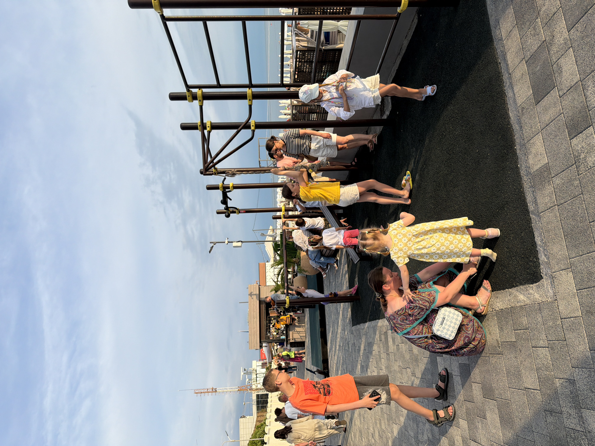 The image depicts a lively outdoor scene, likely in Sochi, Russia, during a late afternoon or early evening. A group of people, predominantly children and adults, are gathered in an open area with a modern playground structure. The playground features dark metal bars with yellow accents, suitable for various physical activities.
In the foreground, a woman in a colorful, patterned dress is kneeling to attend to a young girl in a bright yellow floral dress. The woman has a checkered handbag slung over her shoulder. To the left, a boy in an orange t-shirt and grey shorts stands looking towards the right, holding a smartphone.
The middle ground reveals more activity. Several children are engaged with the playground equipment. A girl in a yellow top and white shorts is holding onto a rope swing. Another girl, wearing a white outfit and a light-colored cap, stands near a metal bar. An adult in a striped shirt and white shorts supervises nearby. Other individuals, including families and children, are visible in the background, some walking and others sitting.
Behind the playground area, there's a glimpse of what appears to be a commercial space or a cafe with people dining or socializing. A tall communication tower is visible in the far distance, with a street lamp nearby. The ground is paved with interlocking grey stones, transitioning to a dark, textured surface around the playground. The sky above is partly cloudy with hues of blue and grey, suggesting either approaching evening or a change in weather. The overall atmosphere is one of casual recreation and community engagement.
