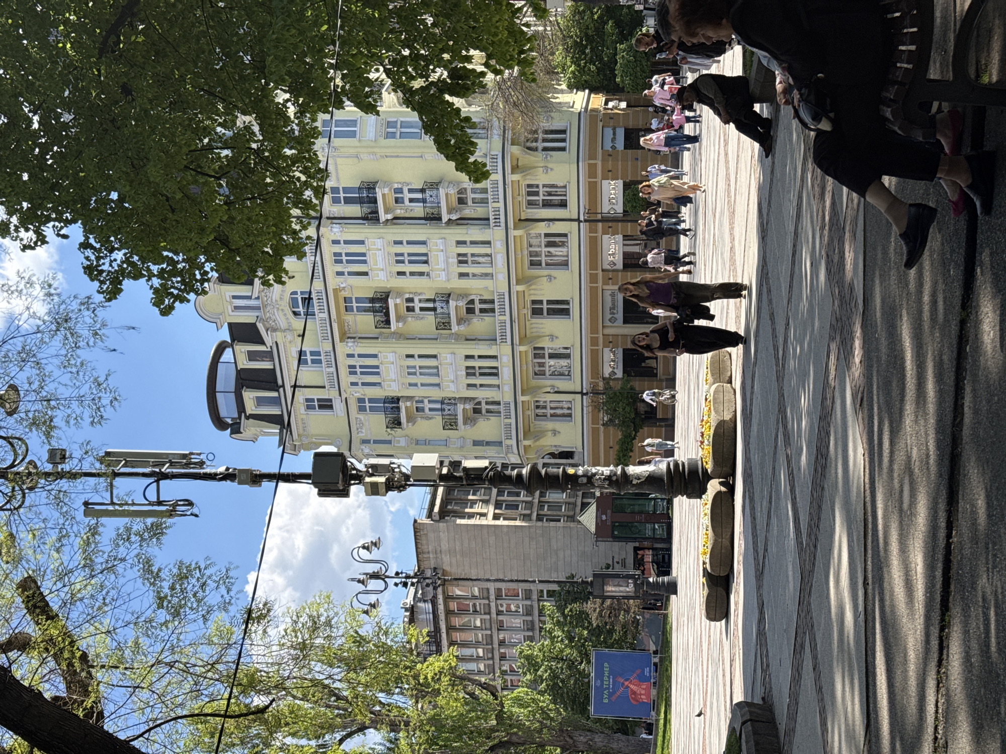 This image shows a street scene in Sofia, Bulgaria. The perspective is from street level, turned vertically. People are walking along a pedestrian area. There are buildings on either side, including a yellow building with many windows. A street lamp with communications equipment is on the left. Trees are also visible along the street. A sign with Bulgarian text is in the foreground. Some people are sitting on benches.