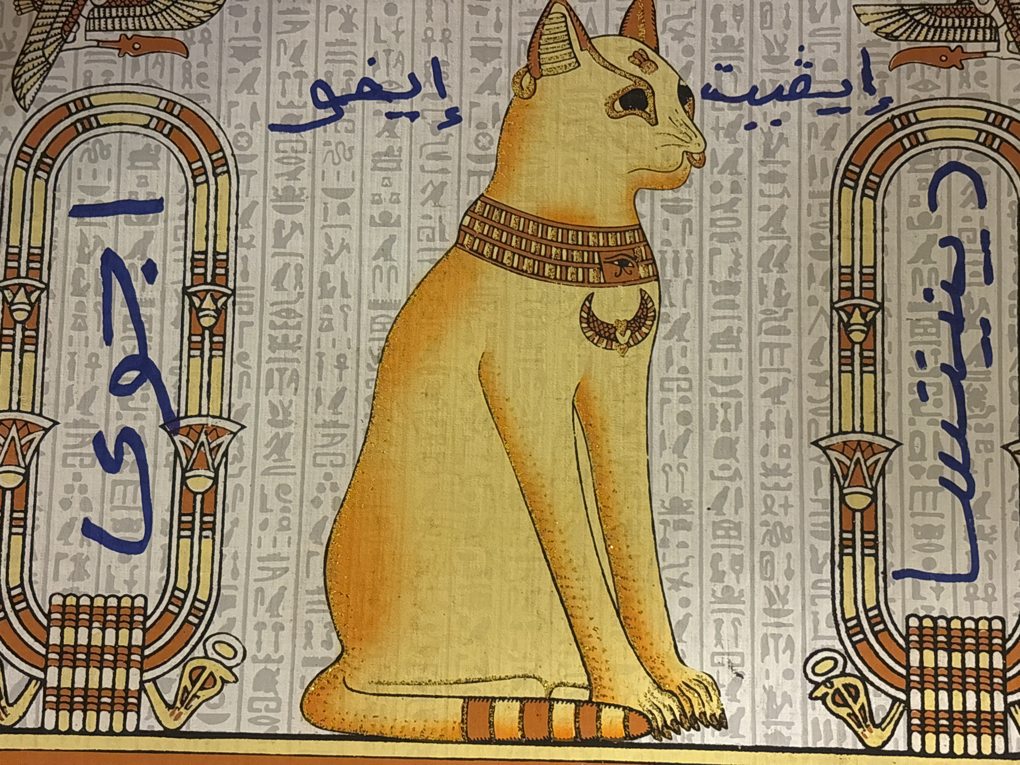 This media file presents a decorative artwork reminiscent of ancient Egyptian papyrus or fresco painting. The central subject is a stylized cat, depicted in profile, sitting regally and facing right. Its fur is a golden yellow, highlighted with sparkling glitter, and it has pointed ears and detailed paws. The cat wears an elaborate collar featuring patterns, an "Eye of Horus" symbol, and a prominent winged crescent moon amulet on its chest.
The background is adorned with columns of various Egyptian hieroglyphs on a light, neutral-toned surface. Flanking the central cat figure are two ornate cartouche-like frames, decorated with striped patterns in orange, yellow, and brown, and featuring stylized floral or lotus motifs at their bases. Partial images of winged figures, possibly representing deities like Horus, are visible at the top edges of the artwork.
Overlaid on this Egyptian-themed background are distinct inscriptions in blue, handwritten-style Arabic script. Inside the left cartouche, the text clearly reads "إخناتون" (Akhenaten). Above the cat's head, to the left, the script says "إيمو" (Imo or Aymo), and to the right, it appears to be "إيقتيت" (Iqtīt or similar). The blue script within the right cartouche is less clearly discernible but follows the same calligraphic style.
The artwork is static, depicting no active event, and serves as a decorative piece combining ancient Egyptian iconography with modern calligraphic elements. The location context for the media file is Sofia, Bulgaria.