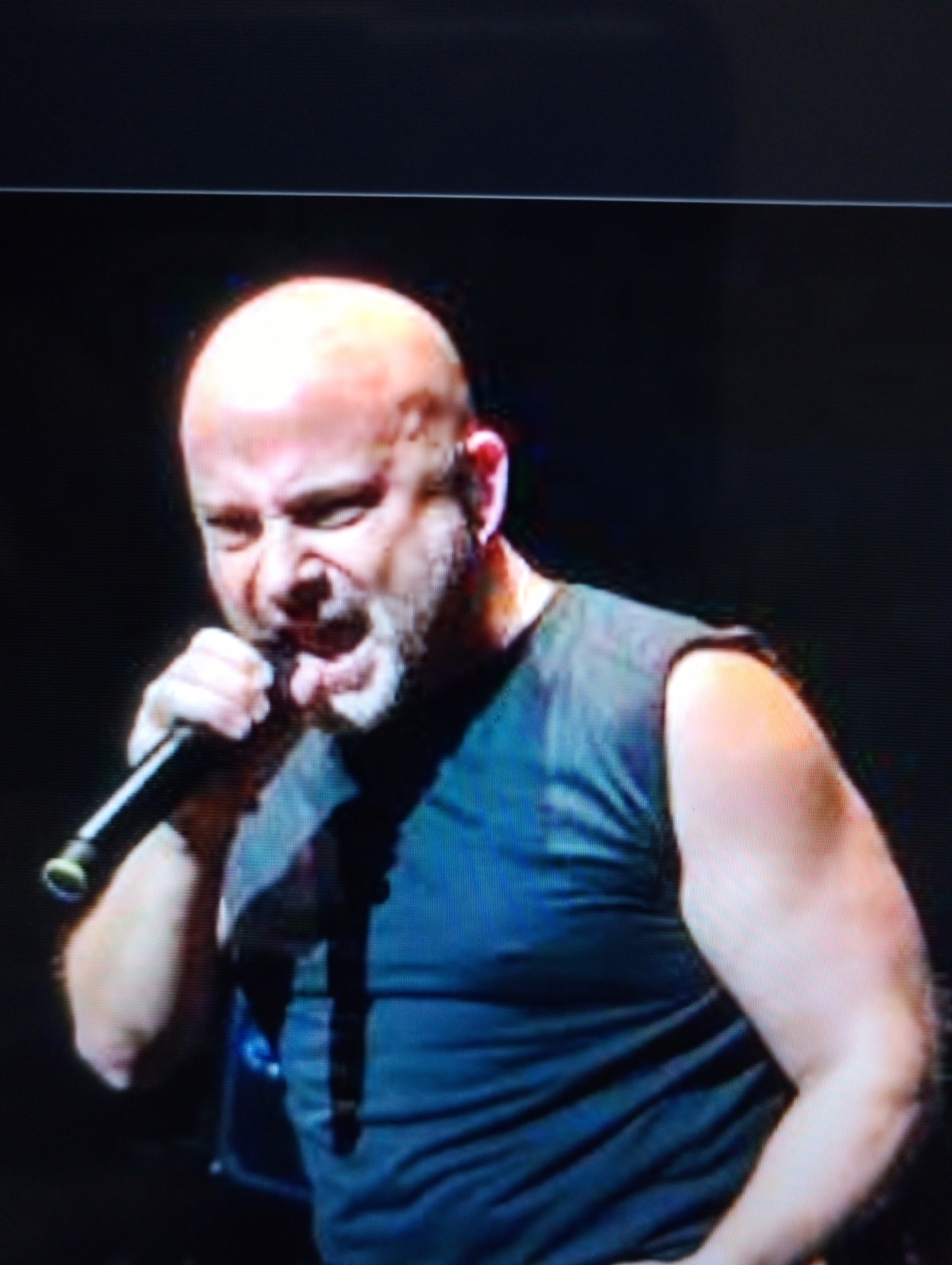 A bald man with a beard is singing into a microphone. He is wearing a dark t-shirt. The background is dark.