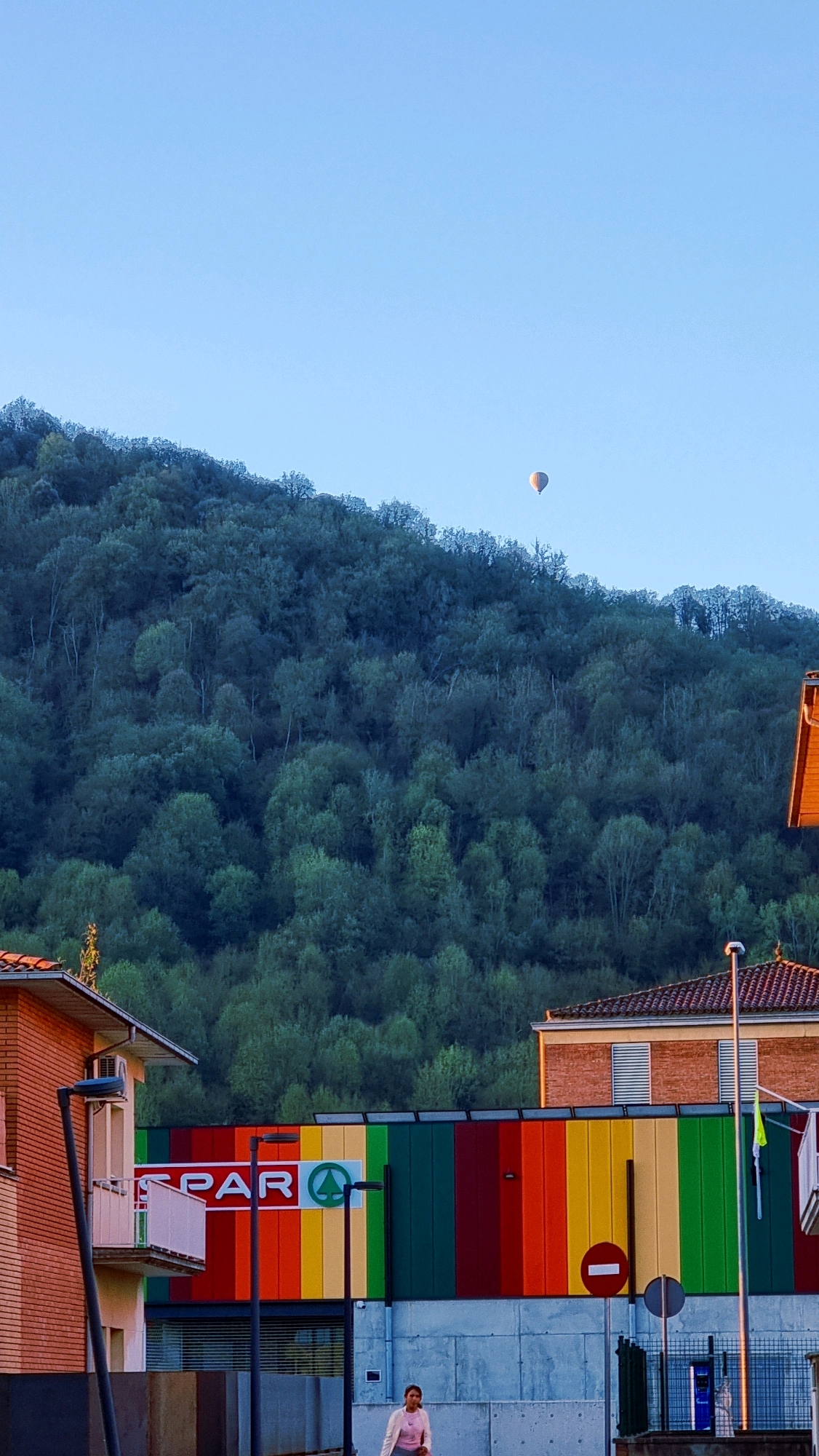 In Sant Joan Les Fonts, Spain, a hot air balloon floats in the clear blue sky above a verdant, tree-covered mountain. Below the mountain, a building with a vibrant, multi-colored facade displays the SPAR logo. A woman walks in front of the building, with a streetlamp and traffic signs also visible. The scene appears to be set in the early morning or late afternoon, with the warm sunlight casting long shadows.