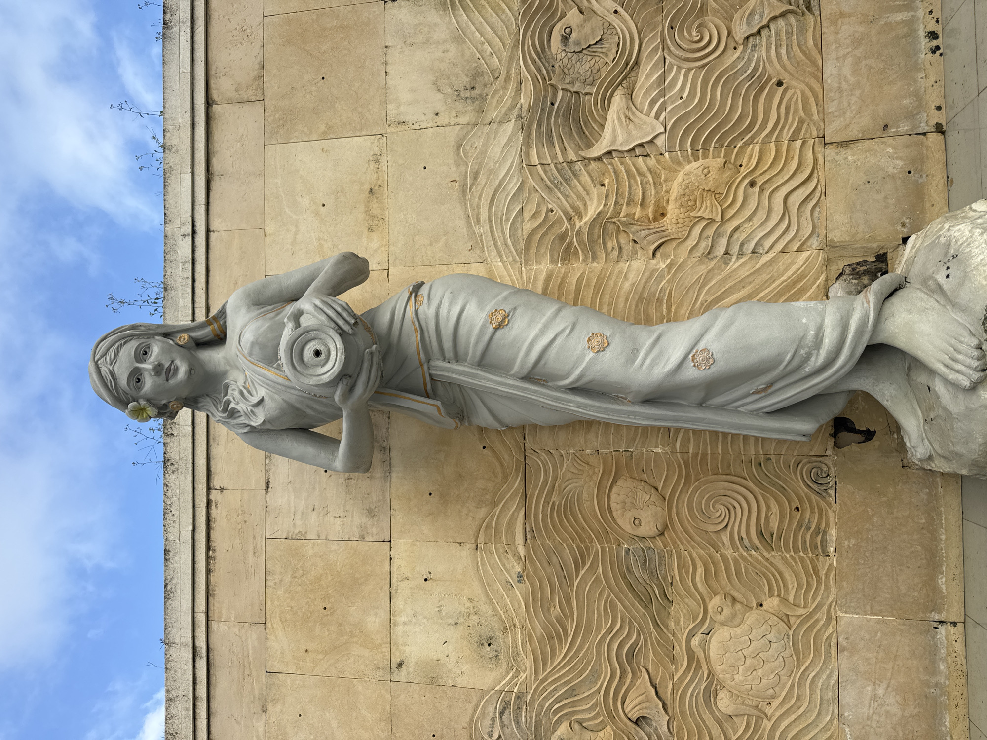 A full-length statue of a woman is affixed to a wall in Jimbaran, Indonesia. The statue is pale grey in color and depicts a woman lying on her side, holding a spherical object to her chest. She is adorned with a flower near her left ear, and gold-colored details are visible on her garment.
The wall behind the statue is made of tan-colored stone blocks, some of which are carved with depictions of fish and swirling water patterns. A narrow strip of blue sky and vegetation is visible above the wall.