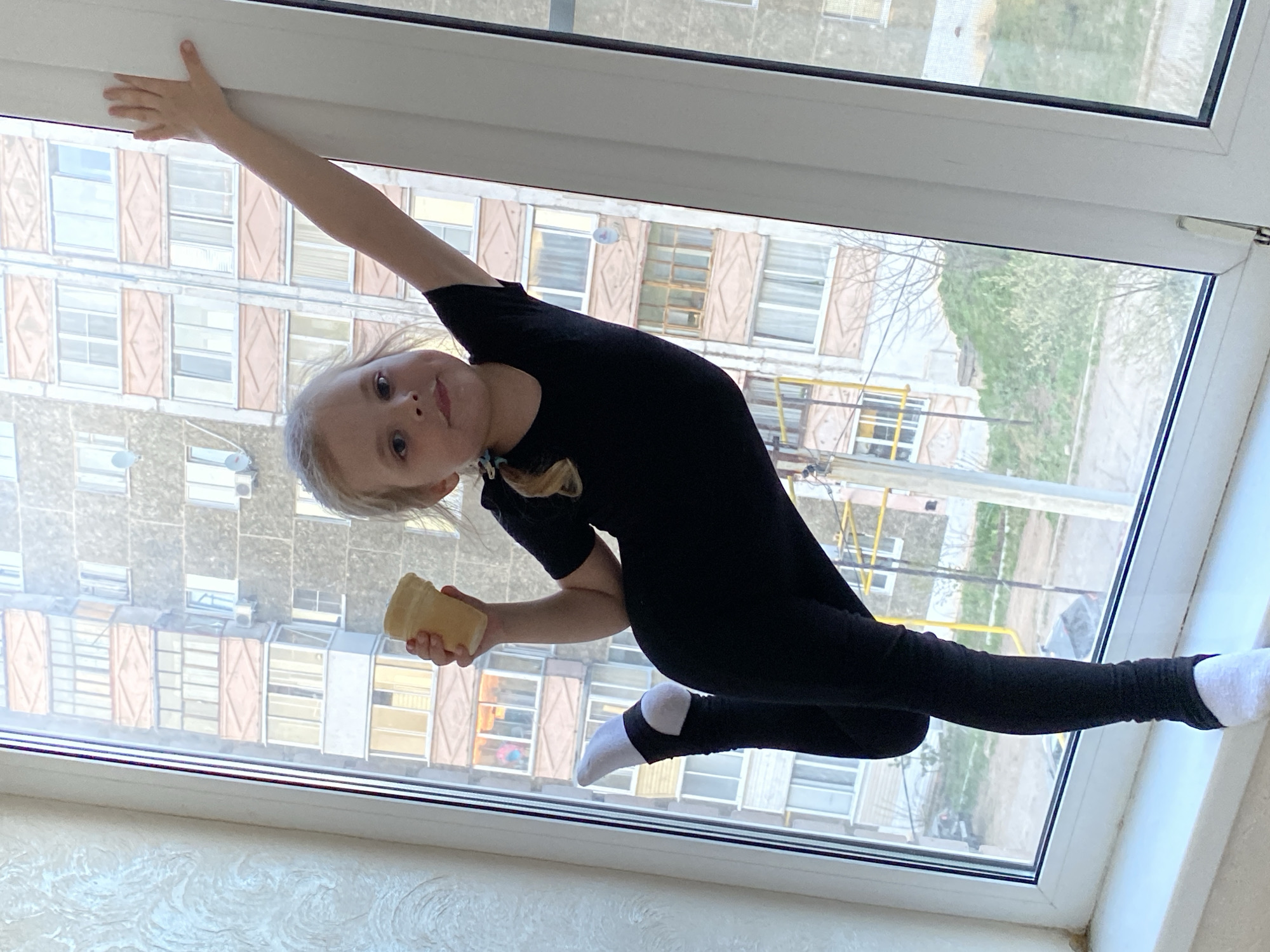 A young child in a black leotard and white socks is positioned precariously on a window sill in Votkinsk, Russia. The child is holding onto the top of the window frame with one hand and balancing on the edge of the sill with one foot. In the other hand, the child is holding a small, beige object. Outside the window is a multi-story apartment building with rows of windows. The sky is overcast, and the overall lighting is subdued.