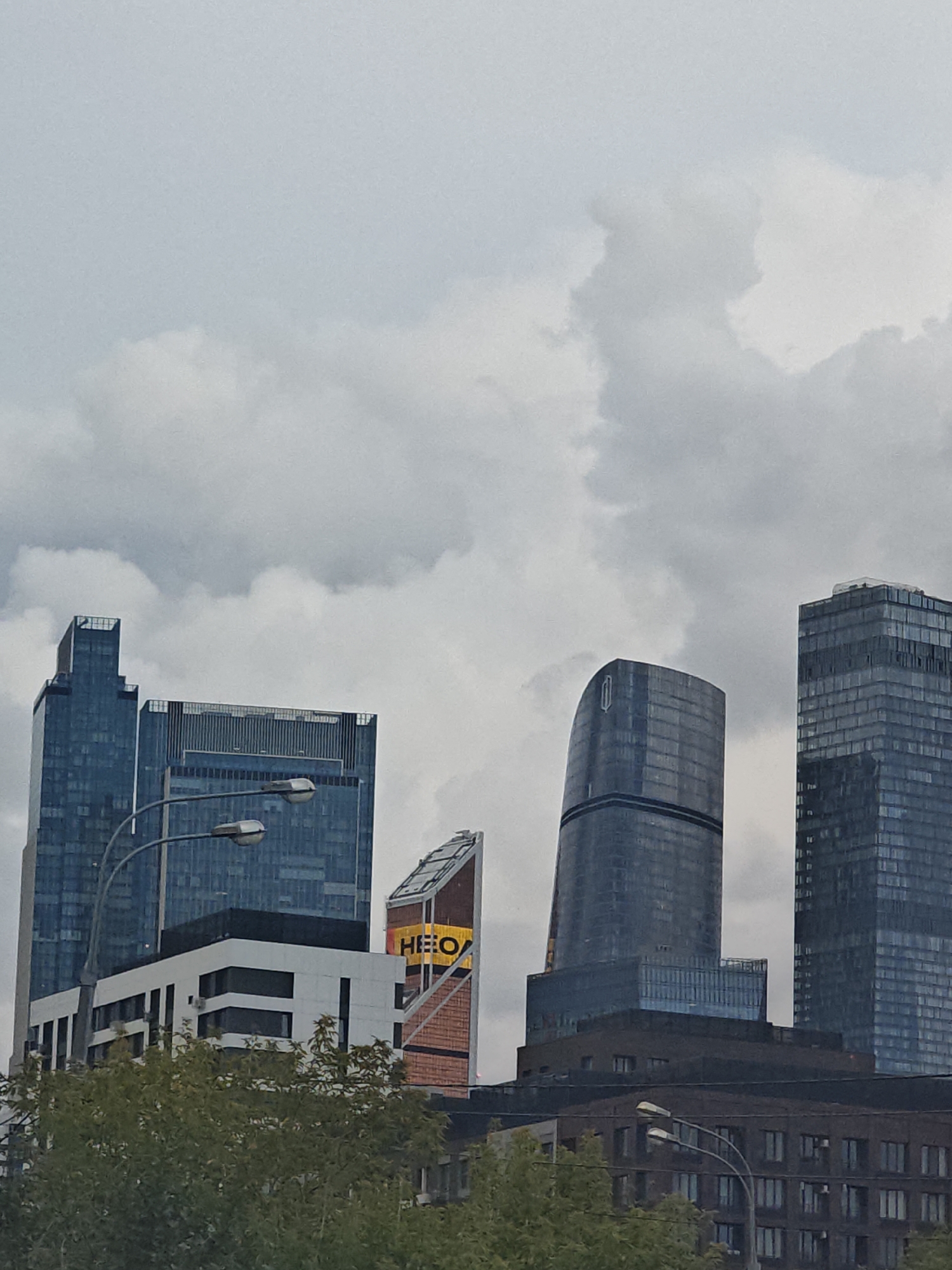 The image shows a cluster of modern skyscrapers in Moscow, Russia. The buildings are characterized by their glass facades and varied architectural styles, with one building featuring a distinctive curved shape. In the foreground, a street lamp with two lights is visible, and trees partially obscure the lower parts of the buildings. The sky is overcast with clouds. There are no people or vehicles visible in the scene. The overall impression is of a business or financial district.
