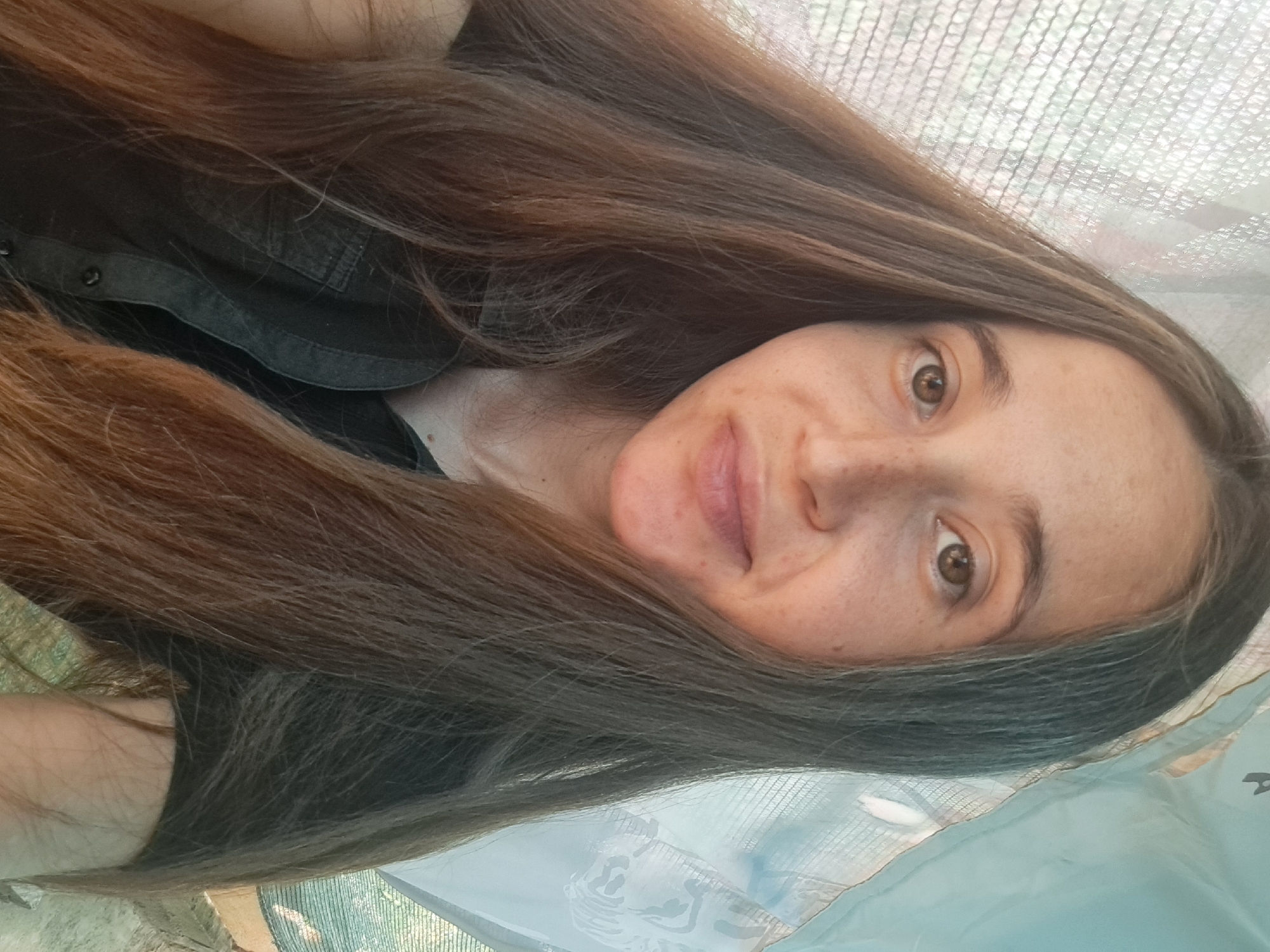 This is a close-up, front-facing selfie of a young woman with long, brown hair and fair skin. She has brown eyes and a light smattering of freckles across her nose and cheeks. She is wearing a dark, collared shirt, possibly denim. The background is out of focus and appears to be outdoors, with a glimpse of a patterned fabric or tarp on the right and a netted structure on the left, suggesting she might be in a garden or under some form of shelter. The lighting suggests it is daytime. The location of Vypasne, Ukraine is not visually discernible from the image itself.