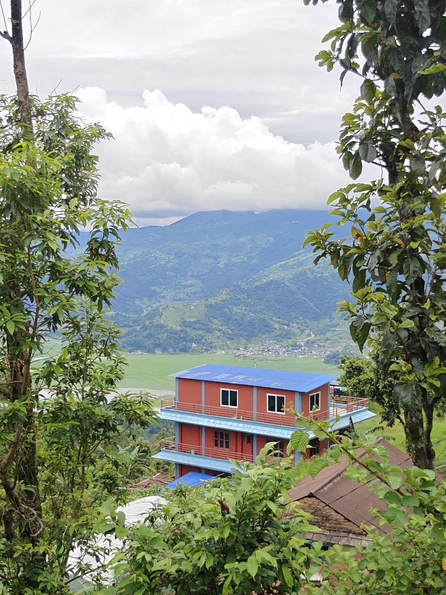 The image depicts a multi-story red building with blue trim and roofing, situated on a hillside overlooking a verdant valley in Pokhara, Nepal. The building has multiple levels with visible windows. In the foreground, lush green trees frame the scene, obscuring parts of the building and the landscape beyond. The valley below shows signs of agricultural activity, with a large, flat green field likely used for rice cultivation. Further in the distance, rolling green hills rise, some of which are terraced, indicating a rural or semi-rural environment. The sky is overcast with scattered clouds, suggesting a diffused light. The overall impression is of a tranquil, natural setting with human habitation integrated into the landscape. The location appears to be a residential area or guesthouse perched on an elevated position, offering a panoramic view of the Pokhara valley and surrounding hills.