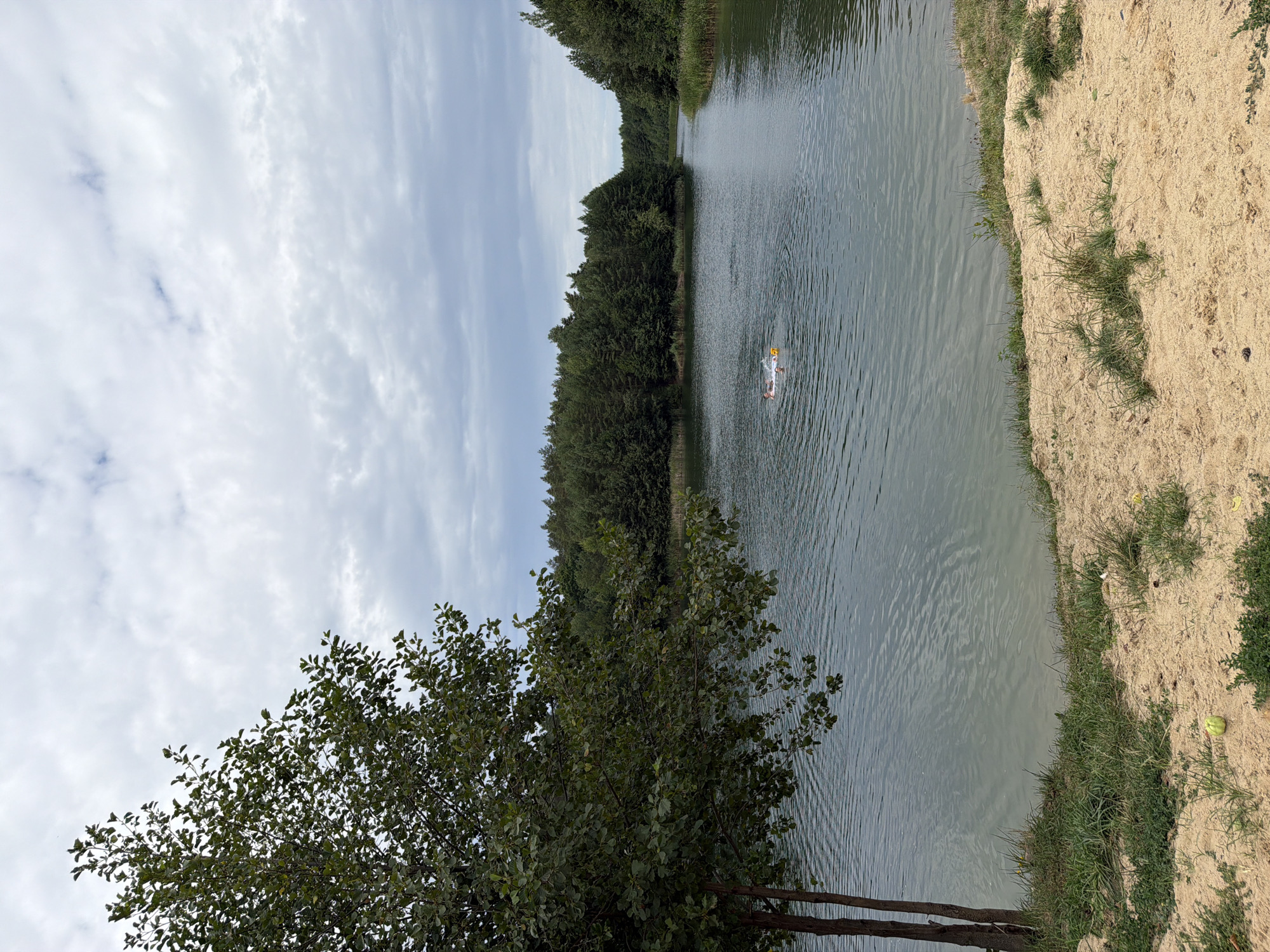 The image depicts a tranquil scene at a lake located in Kmytiv, Ukraine. In the foreground, a sandy embankment with sparse green grass slopes down towards the water. A tree with dense green foliage stands on the left side of the embankment, its branches extending over the water. The lake itself is calm with ripples on the surface, reflecting the overcast sky. In the middle of the lake, a pink inflatable raft is visible with at least two individuals on it, one of whom appears to be holding a yellow object. The lake is surrounded by a dense forest of green trees, with a narrow channel of water leading further into the distance. The overall atmosphere is serene and natural.