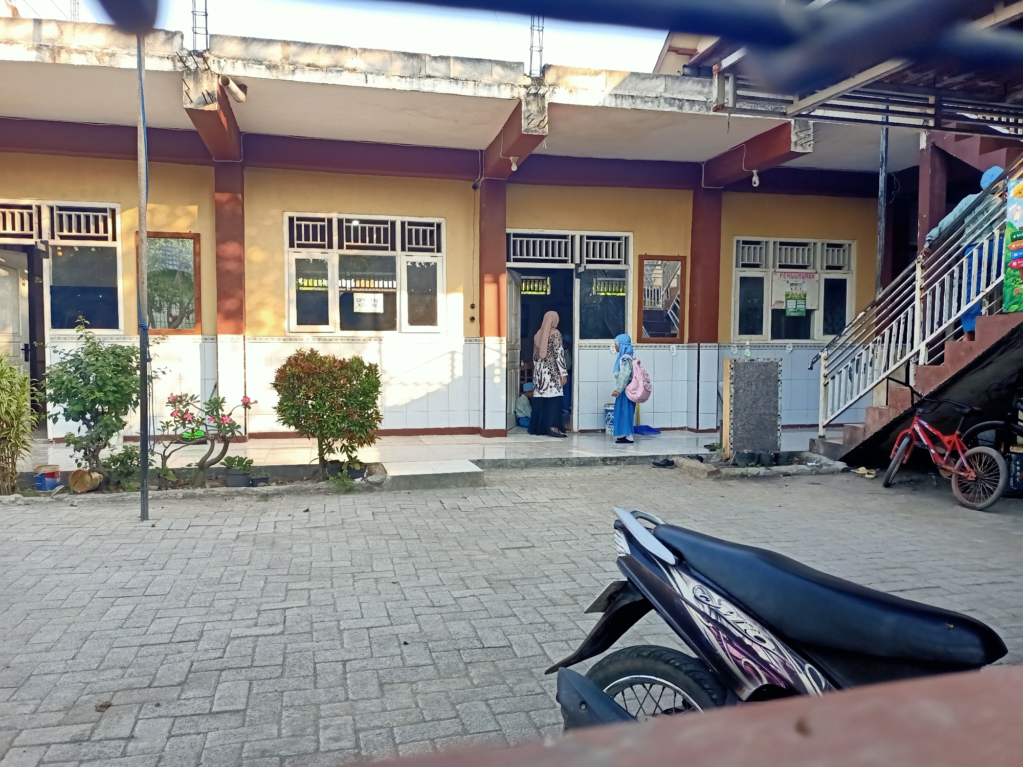 The image depicts the exterior of a building with a covered walkway, possibly a school or administrative office, in Banjarjo, Indonesia. The building features a yellow facade with brown trim and white tiled wainscoting. Several windows with white bars are visible. In front of the building, a paved area with interlocking bricks is present, with a few plants and trees in planters.
Two women, dressed in modest clothing and headscarves, are near an entrance. One woman in a patterned dress is standing, and another in a blue outfit is carrying a pink backpack and walking away from the entrance. Further to the right, a red bicycle is leaning against the building near a set of outdoor stairs. A motorcycle is parked in the foreground, partially obscuring the view of the paved area. The overall environment suggests a daytime scene with bright sunlight casting shadows. The building appears to be a single-story structure with an upper level visible in the background on the right. A mirror hangs on the wall near one of the entrances, reflecting trees and the sky.