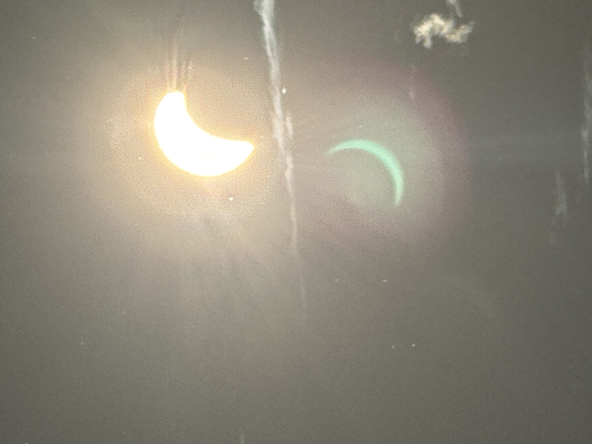 The image shows a solar eclipse, with a crescent-shaped sun visible against a hazy, gray sky. A reflection or secondary image of the crescent sun, tinted green, is visible below the primary image. Wispy clouds are present in the sky. The scene is not identifiable as being in Waterford, Ireland, or any specific city.