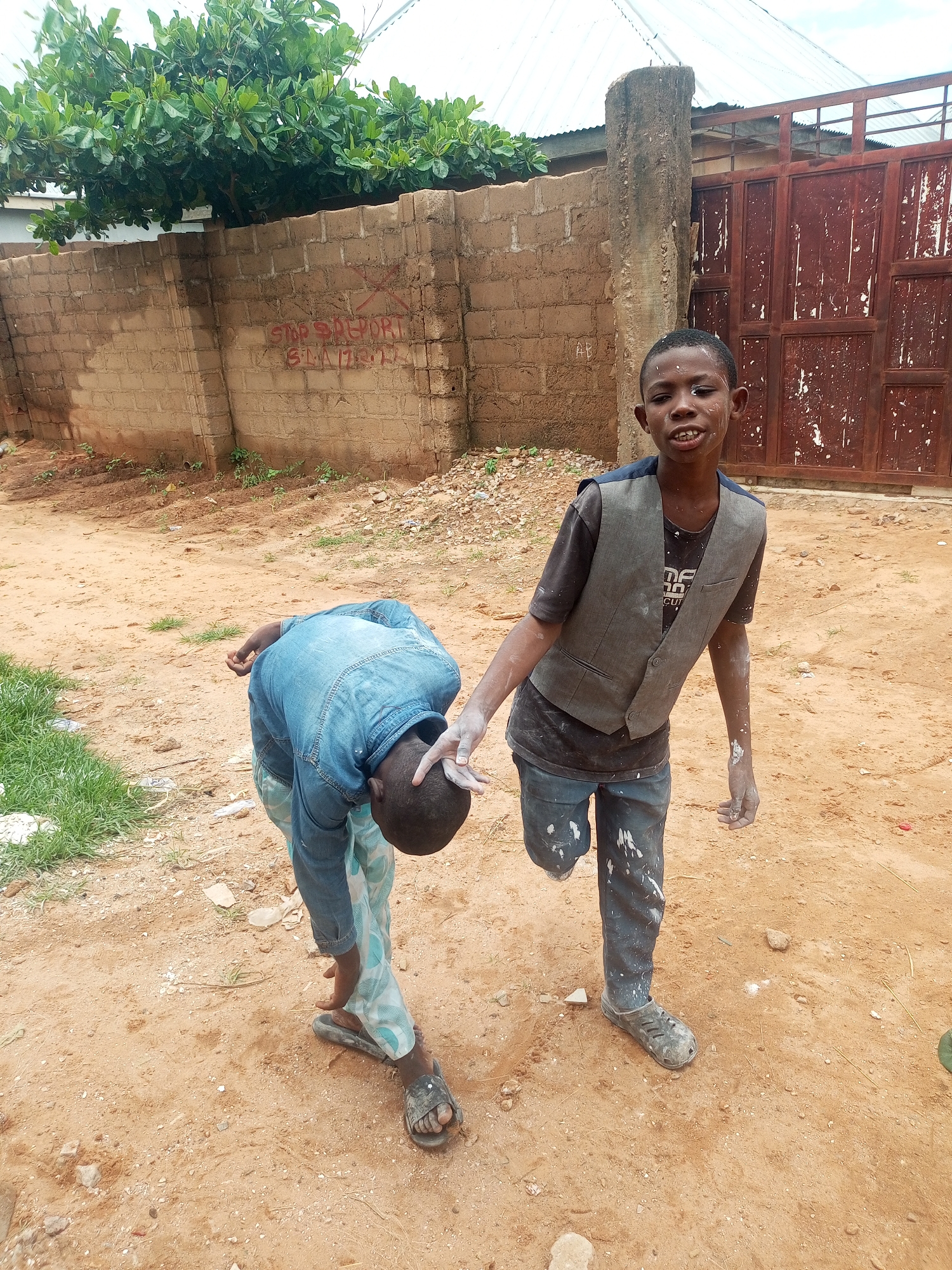 In the city of Tempure, Nigeria, two young boys are present in an outdoor environment. One boy, with a prosthetic right leg, stands facing the camera and appears to be looking to his left. He wears a dark t-shirt, a grey vest, and dark trousers that are splattered with white material. He has white powder or paint on his face, hands, and arms. The second boy is bent over in front of the first, facing away from the camera. He is wearing a blue denim shirt and patterned trousers. His right hand is visible, extended towards the standing boy's left hand, and also appears to have white powder on it. They are standing on a dirt ground with scattered debris and small green plants growing in patches. Behind them is a brick wall with red graffiti that reads "STOP SPELLING SL..." and a metal gate. A large tree with green foliage is visible above the wall. The sky is overcast.