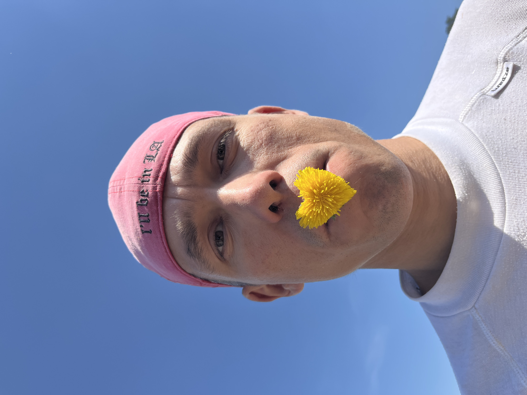 A man with a pink hat and a yellow dandelion flower in his mouth is pictured from below against a bright blue sky. He wears a white shirt with "Uredze" printed on the collar. The hat has "I'll be in LA" printed on it. The location appears to be Chaikovsky, Russia, based on the possible reference to the Uredze brand.