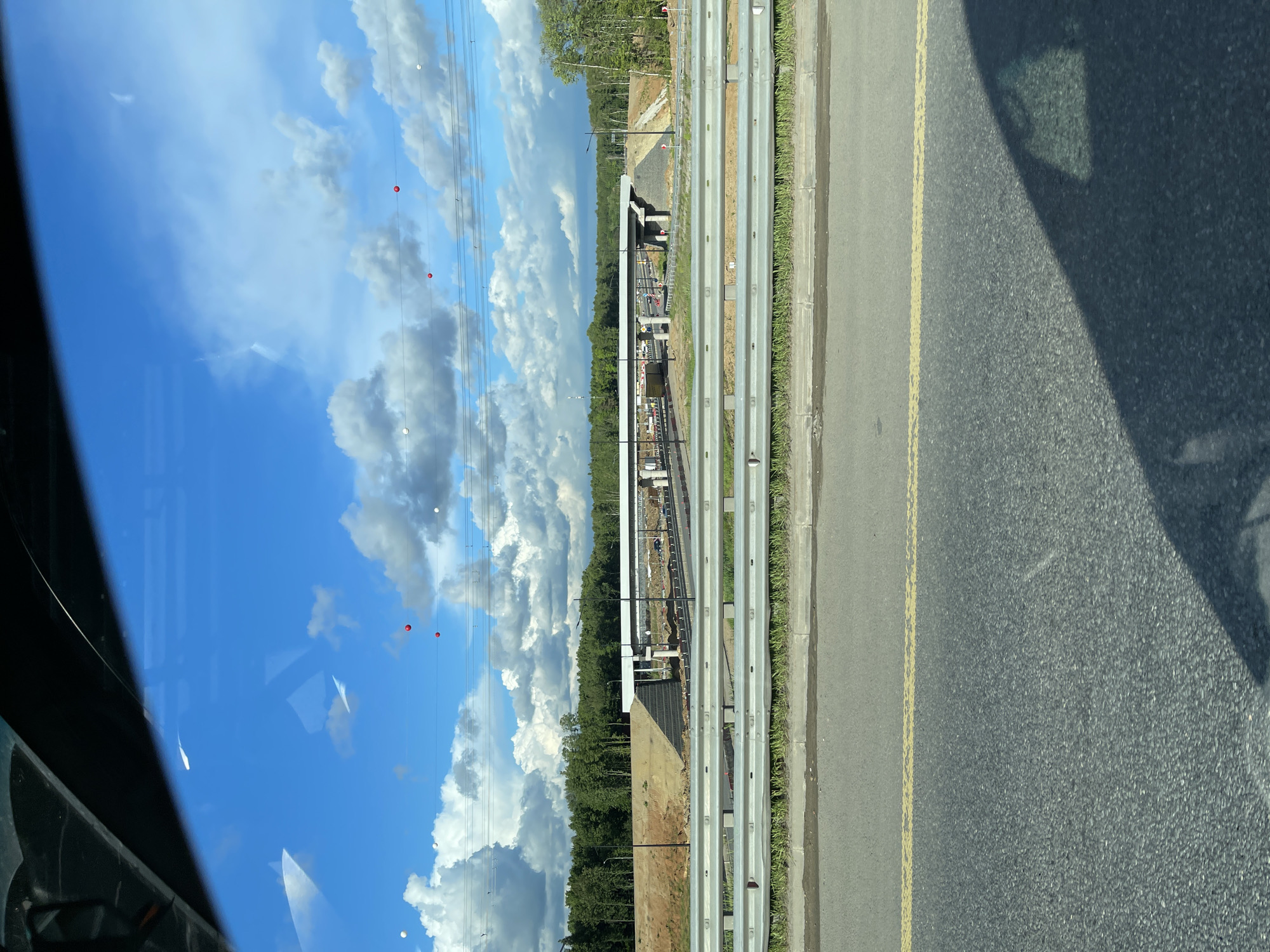 The image shows a view from inside a vehicle, possibly a car, driving on a road in Novodrozhzhino, Russia. The road has a double yellow line marking, a concrete curb, and what appear to be guardrails. Beyond the guardrails is a construction site with heavy equipment visible. The sky is blue with scattered white clouds. Reflections are visible on the vehicle's window. Overhead power lines with red and white markers are present. The image is rotated 90 degrees clockwise.