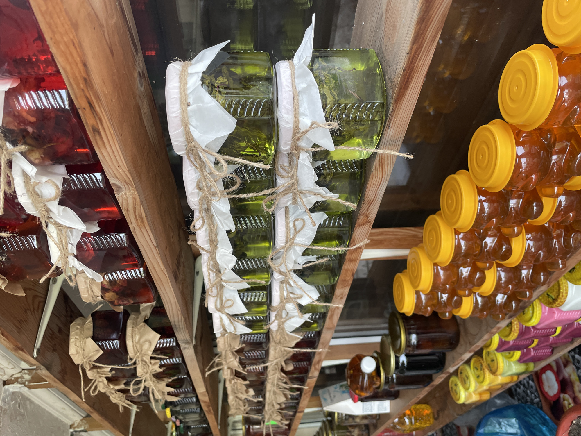 The image depicts a low-angle, eye-level view of shelves stocked with jars of preserves, liqueurs, and honey. The scene appears to be inside a storage space or shed. Jars filled with a red liquid line the left side of the frame, while jars filled with a green liquid, wrapped in twine and white paper, sit in the center. To the right, jars of honey with yellow lids line the shelf. Various other jars, including some with yellow lids and purple wrapping, can be seen on the bottom right. The shelves are made of rough, unfinished wood. The image was taken indoors, but there is natural light coming through a window in the background. The location of the image is Olginka, Russia.