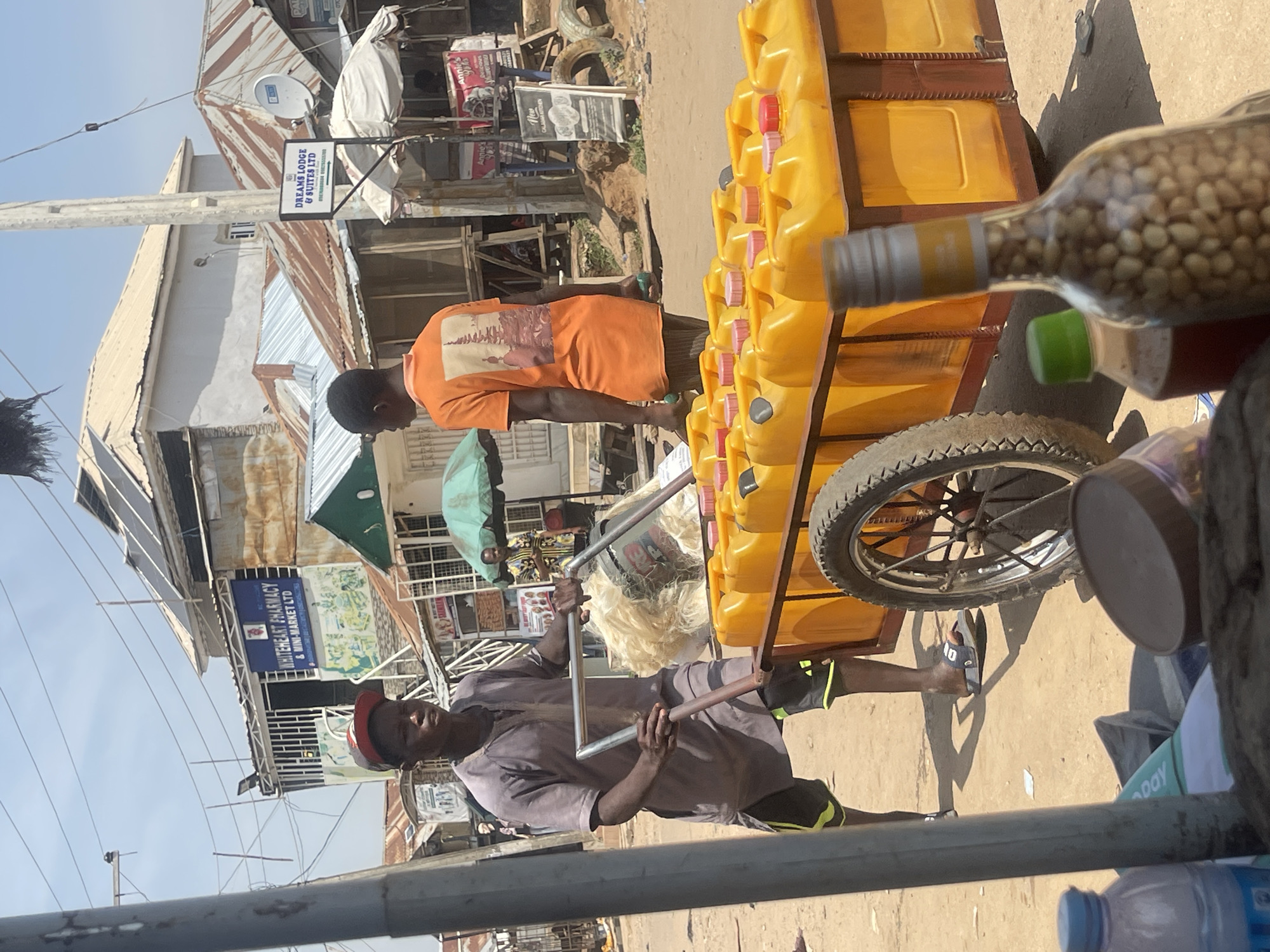 This is a medium shot of a street scene in Abuja, Nigeria, under a bright, sunny sky. In the foreground, a young man, wearing a gray tunic, dark shorts with neon green trim, and a red baseball cap, pushes a cart laden with yellow plastic jerrycans. The cart has a single wheel and appears to be made of metal. He is holding onto the silver metal handles of the cart. Another young man, wearing an orange t-shirt with a graphic print on the back, stands to the right, facing away from the camera. In the background, there are several buildings with corrugated iron roofs, typical of informal settlements or older commercial areas. One building displays a sign that reads "WHITEHEART PHARMACY & MINI-MARKET LTD". Another sign visible reads "DREAMS LODGE & SUITES LTD". The ground is dusty and uneven, with scattered debris. A bottle filled with small, light-colored objects, possibly nuts or seeds, is in the immediate foreground, obscuring some of the cart and the ground. To the left of the bottle is another smaller bottle with a green cap and a darker liquid. The overall atmosphere suggests a bustling marketplace or commercial street.