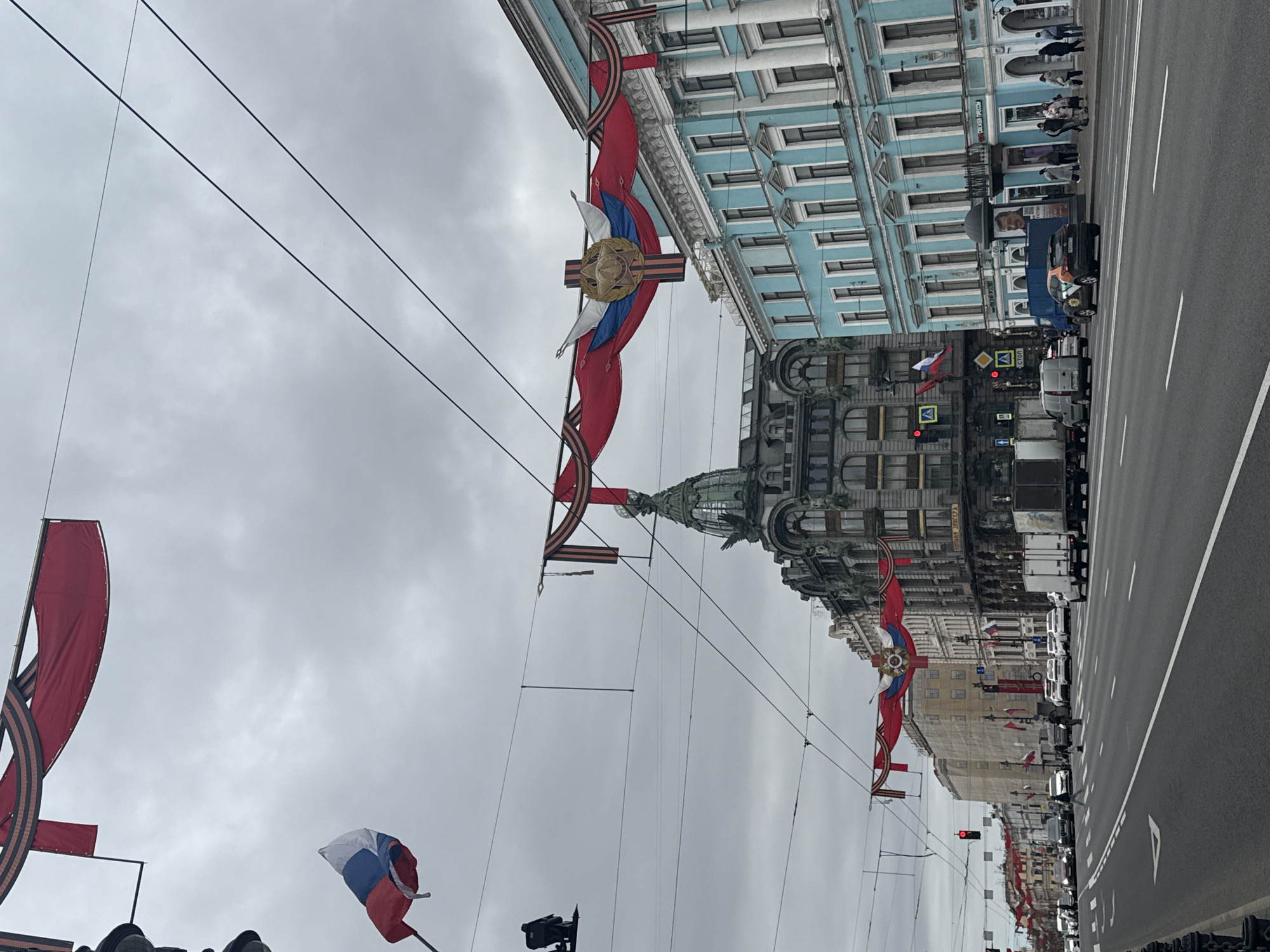 This image captures a vibrant street scene in Saint Petersburg, Russia, adorned with celebratory decorations under an overcast sky. The view looks down a wide asphalt road, flanked by multi-story buildings of varying architectural styles.
Prominently displayed across the street are large, colorful banners and ribbons. These decorations feature the red, white, and blue of the Russian flag, often intertwined with the distinctive black and orange St. George's ribbon. Many decorations also incorporate a gold star emblem, frequently associated with military valor, suggesting a commemoration like Victory Day. Smaller Russian flags are also visible, some attached to poles and others flying independently. Overhead, numerous wires crisscross the sky, likely for public transport like trams or trolleybuses.
On the right side of the street, a light blue building with numerous windows and classical architectural elements stands out. Across the street, a more ornate, darker building with a distinctive green dome and intricate facade is visible. This iconic structure is identifiable as the Singer House (also known as the House of Books).
The street itself is busy with several cars and trucks, some parked and others in motion, indicating active city life. Pedestrians can be seen on the sidewalks. The sky is uniformly grey and cloudy, suggesting an overcast day rather than bright sunshine.
Visible text on the buildings includes Cyrillic signage. On the light blue building, parts of words like "БЕЛЫЙ ГРИФОН" (White Gryphon) are discernible, and further down, another sign shows "СО...ТЕЛЬ". On the darker, ornate building, the sign clearly reads "ДОМ КНИГИ" (House of Books). The overall scene conveys a sense of public celebration or commemoration in a historical urban setting.