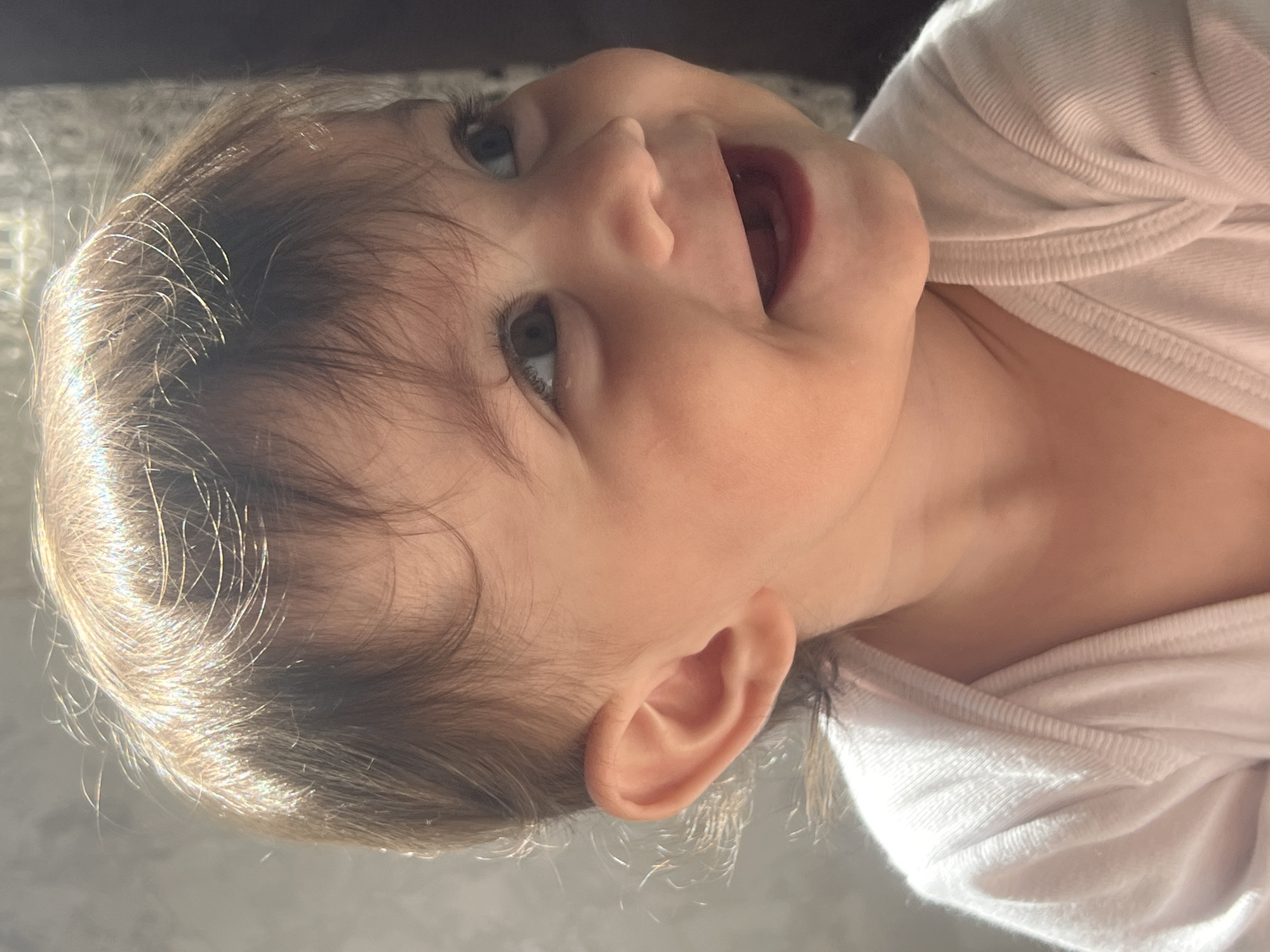This close-up portrait, taken in Elena, Bulgaria, features a young child, likely a toddler, captured with a joyous and open-mouthed smile as they look upwards. The child has light brown hair, which is strongly highlighted by natural light, suggesting sunlight from a nearby window, possibly in the morning or afternoon. Their dark eyes are expressive, reflecting the light. The child is wearing a plain white, ribbed garment, visible around their neck and upper torso. The background is softly blurred, indicating an indoor setting but without specific details. The overall impression is one of happiness and contentment. There is no visible text in the image.