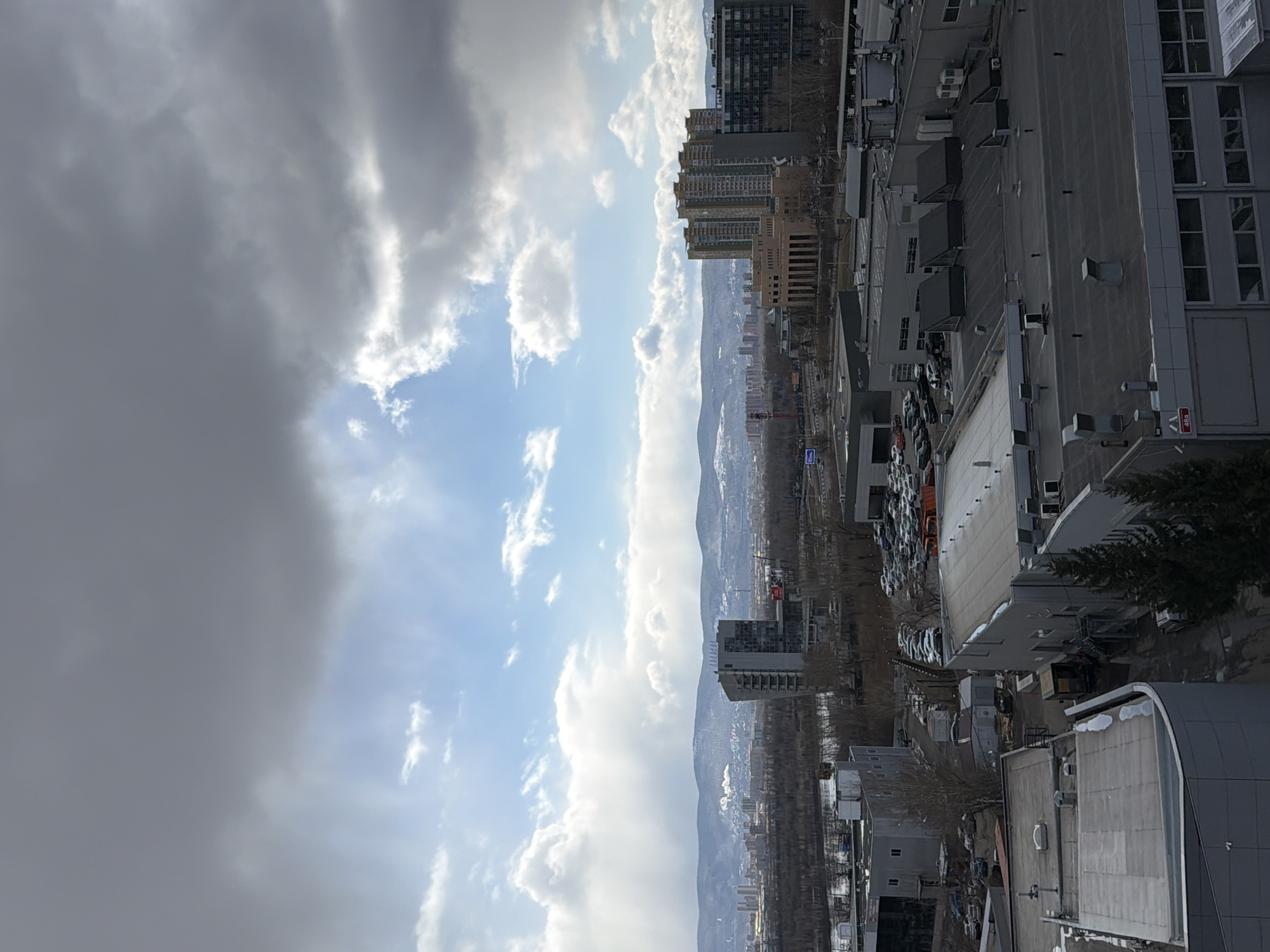The image depicts an elevated view of Krasnoyarsk, Russia. The sky dominates the left side, with dark storm clouds contrasting with patches of blue sky and white cumulus clouds. The city stretches out to the right, nestled against a backdrop of distant mountains. Buildings of varying heights are visible, ranging from low-rise structures to tall apartment blocks. A mix of residential and commercial buildings can be seen, alongside a large parking lot filled with cars. Trees are interspersed throughout the urban landscape, some appearing bare, indicative of late winter or early spring. The overall impression is a city partially obscured by an approaching storm.