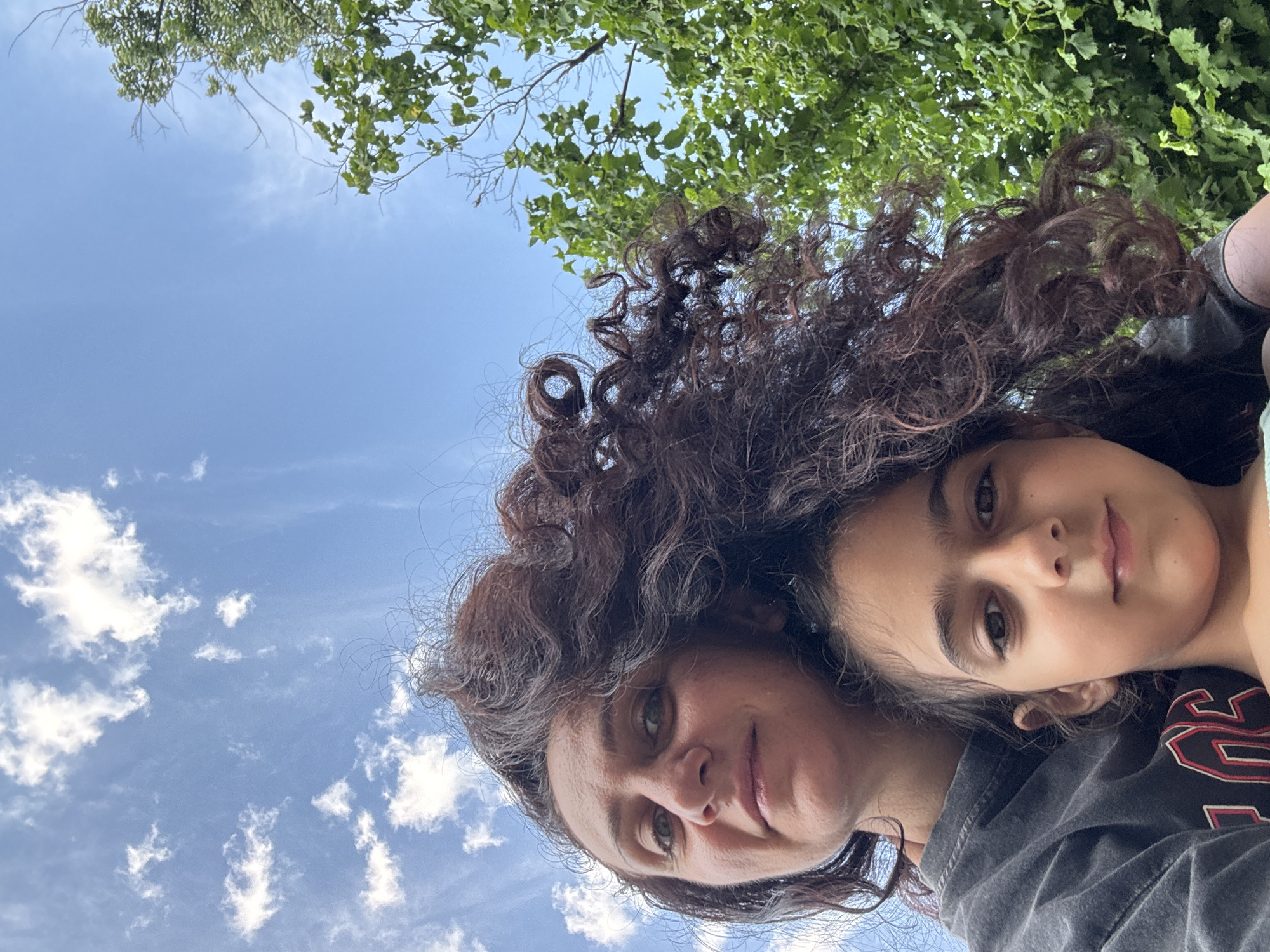 A low-angle, outdoor selfie features a woman and a child with a bright blue sky and green foliage in the background. The woman, positioned on the left, has long, dark, curly hair that dominates the upper portion of the frame. She is looking upwards and to the left with a slight smile. The child is in front of her, with their face visible. The child has dark hair and is looking directly at the camera. They are both wearing casual clothing. The scene appears to be in a park or garden setting, possibly in Sochi, Russia, given the lush greenery and clear sky.