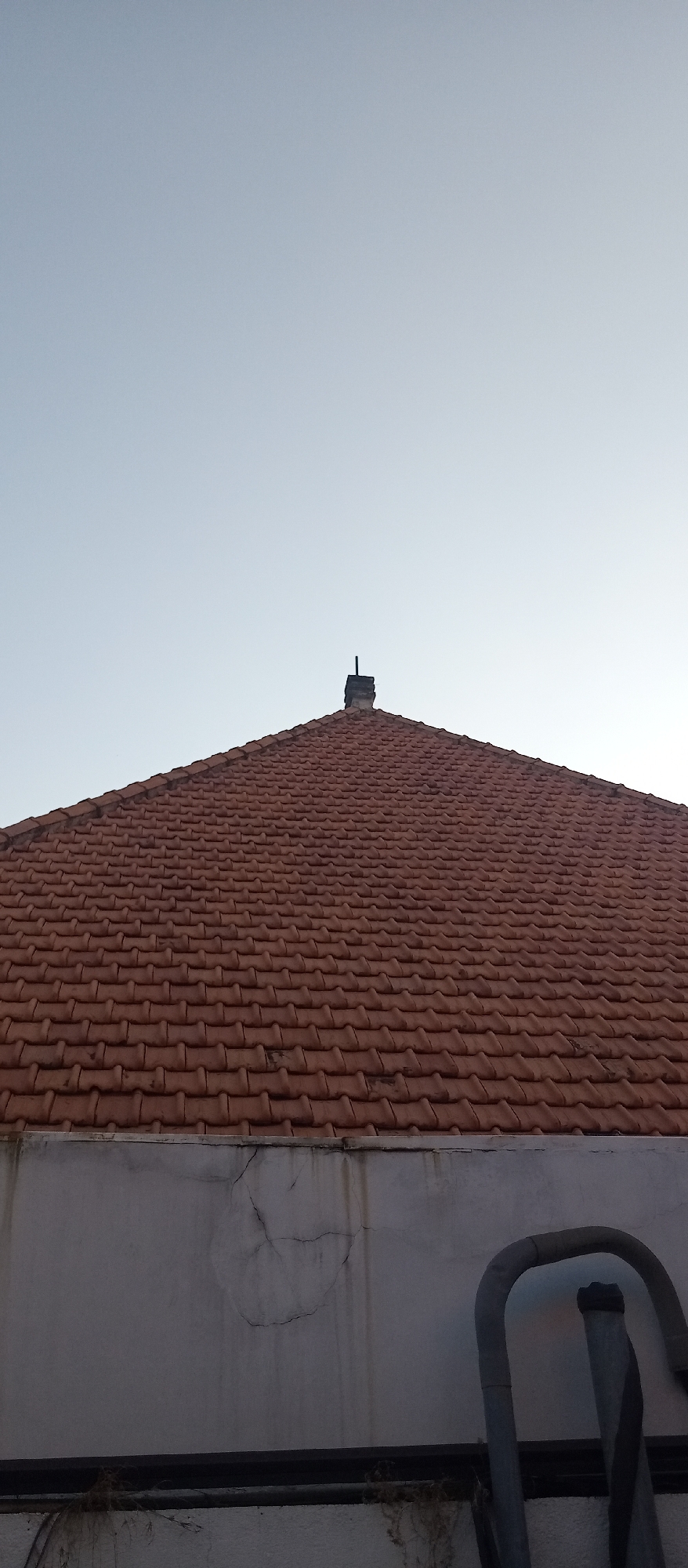 A low-angle shot captures the top portion of a building under a clear, pale blue sky. The dominant feature is a steeply pitched roof covered in reddish-brown, interlocking tiles. A dark, rectangular chimney or vent protrudes from the apex of the roof. Below the roofline, a section of a light-colored, textured wall is visible, showing signs of weathering, including hairline cracks and darker streaks. In the lower right corner, a portion of a curved, metallic structure, possibly part of a ladder or scaffolding, is present. The scene suggests an outdoor urban environment, consistent with a residential or commercial building in Surabaya, Indonesia. There are no people or animals in the frame.