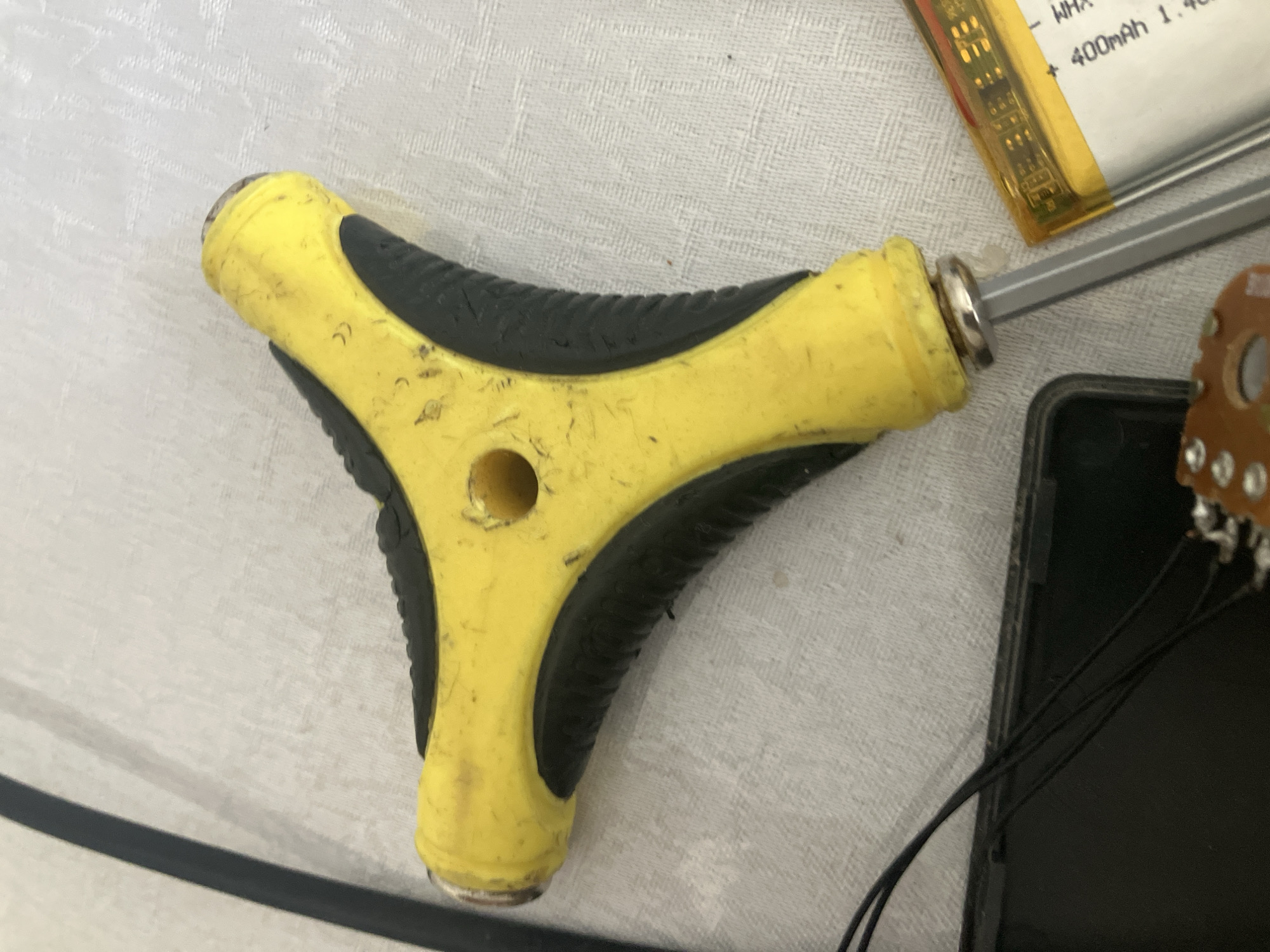 The image depicts a close-up, overhead view of a yellow and black T-handle hex wrench resting on a light-colored, textured surface. The wrench is positioned diagonally across the frame, with the handle towards the top right and the shaft extending downwards and to the left. The yellow plastic handle shows signs of wear, with scuff marks and dirt visible. A circular hole is present in the center of the handle. The black rubber grip sections on the handle have a ribbed texture. A portion of a black cable or wire is visible to the left of the wrench. In the bottom right corner, a yellow object, possibly a battery pack, is partially visible. Text on this object includes "400mAh 1.4". The scene is indoors, and the lighting is adequate. There is no indication of the location being in Tashkent, Uzbekistan, or any specific city. The image focuses solely on the described objects.