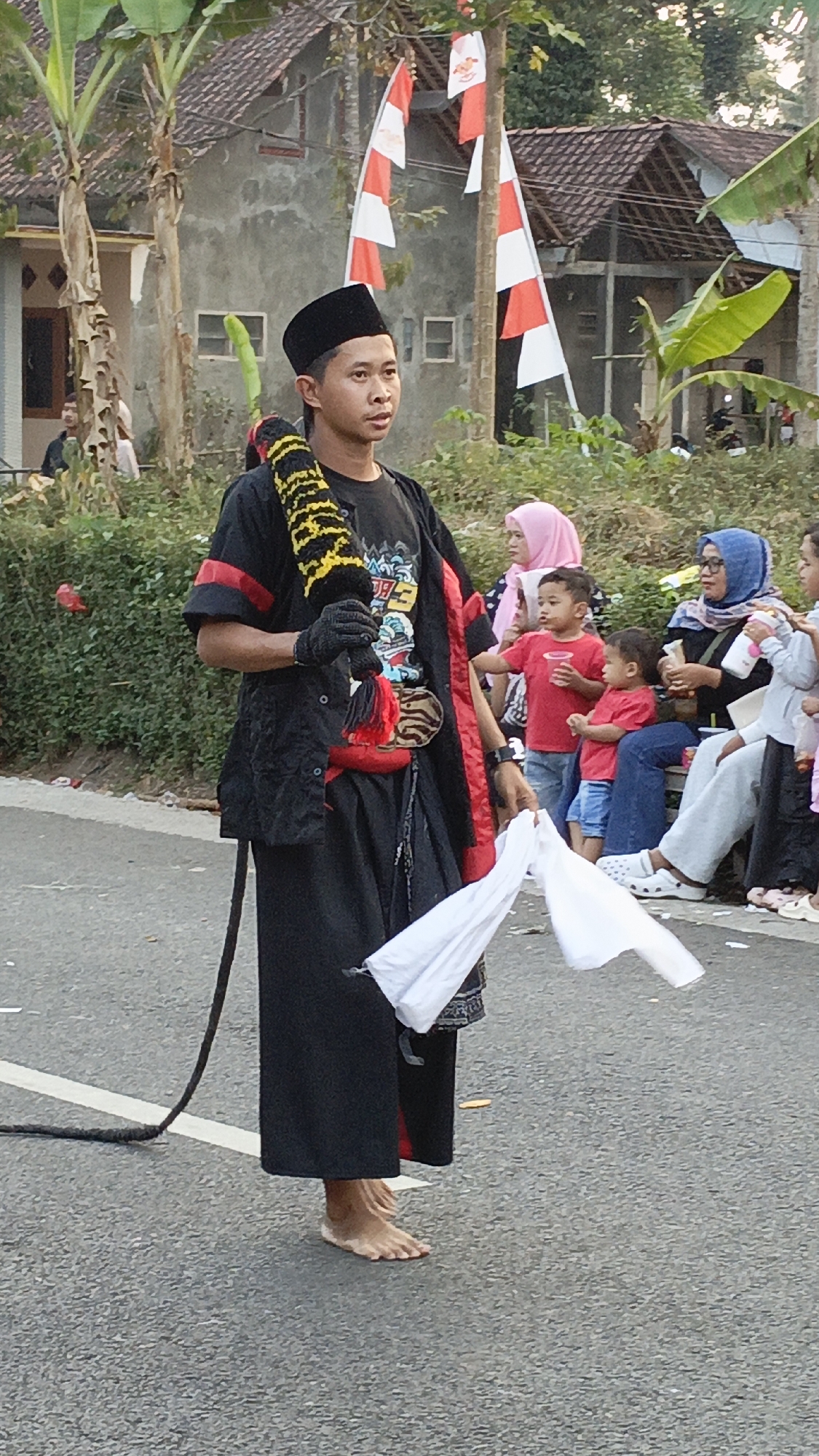 In Krajan Lor, Indonesia, a barefoot young man wearing a black peci, black t-shirt with a graphic print, and a black robe with red trim, stands in the middle of a paved street. He holds a black and yellow braided rope or whip in his right hand, and a white cloth in his left. He is also wearing black gloves and has a red sash around his waist. Behind him, a group of spectators, including children and adults wearing headscarves, observe the scene. Red and white flags are visible in the background, strung on poles. The environment suggests an outdoor event or performance, possibly a parade or cultural celebration, with houses and vegetation visible in the background.