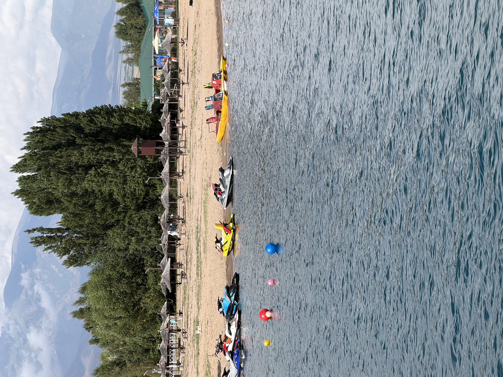 The image depicts a resort area with a beach, trees, and mountains in the background. In the foreground, the blue water of a lake or sea is visible, with several buoys floating on the surface. Along the shoreline, there are several jet skis parked on the sandy beach. Further back, there are numerous straw umbrellas providing shade, and some recreational structures. In the distance, tall green trees form a dense line, beyond which are hazy mountains under a cloudy sky. The scene appears to be a leisure destination, possibly a beach resort or park.