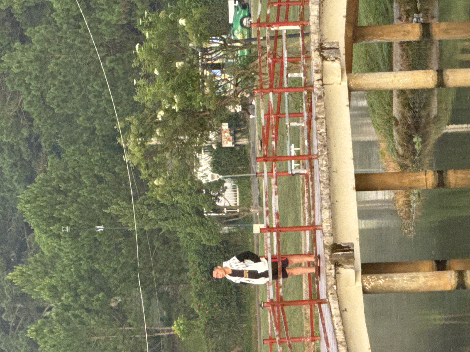 The image depicts a young man standing on a bridge over a body of water, likely a lake or pond. The bridge has red railings. The man is wearing a white hooded jacket with black sleeves and a black t-shirt, along with black shorts and sandals. He is looking to his left. Behind him, on the bank of the water, is a line of trees and some structures, including what appears to be a lamp post. In the background, there are more trees forming a dense forest. A green car is visible further in the background. The overall atmosphere suggests a natural park or recreational area within Phường Xuân Hương - Đà Lạt, Vietnam.