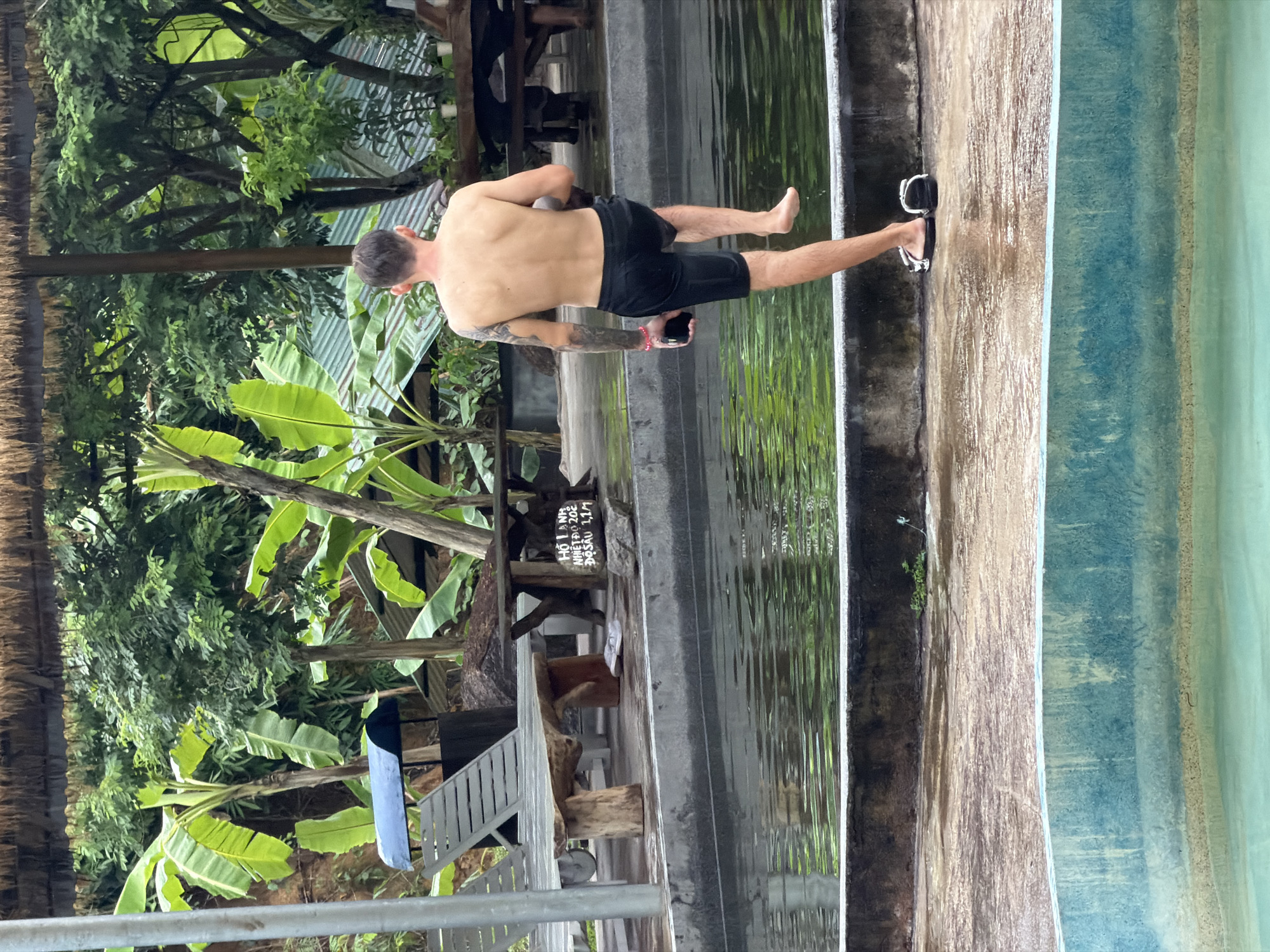 The image depicts an outdoor scene in Khanh Vinh, Vietnam, likely at a pool or water park. A shirtless man, wearing black shorts and flip-flops, is captured mid-stride as he walks along the edge of a pool. He is holding a mobile phone in his left hand and has tattoos on his left arm. The pool's water is a vibrant green, reflecting the lush vegetation surrounding the area, including banana trees and other dense foliage. In the background, there are wooden tables and chairs, as well as lounge chairs, suggesting a recreational setting. The ground around the pool is wet and appears to be a paved surface. A sign with Vietnamese text is partially visible on a wooden table in the middle ground. The overall environment is tropical and humid.