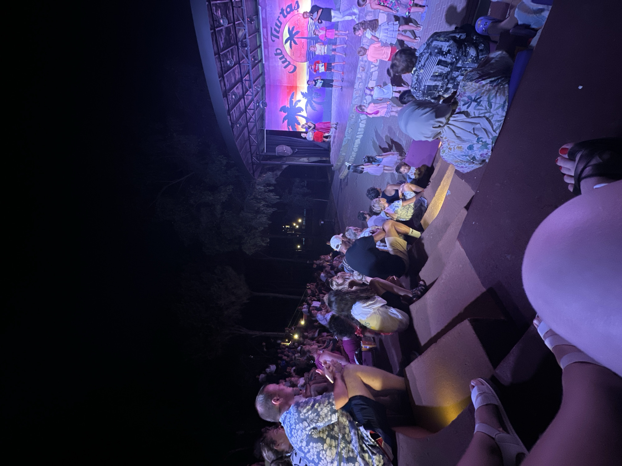 In Alanya, Turkey, an outdoor amphitheater stage is lit for a performance. A backdrop depicts a sunset with palm trees and the words "Club Tartas". Several children and adults are on stage. An audience is seated on tiered, dark red benches facing the stage. The scene is captured at night, with purple and pink lights illuminating the stage. Some audience members are in focus in the foreground, while others are further back.