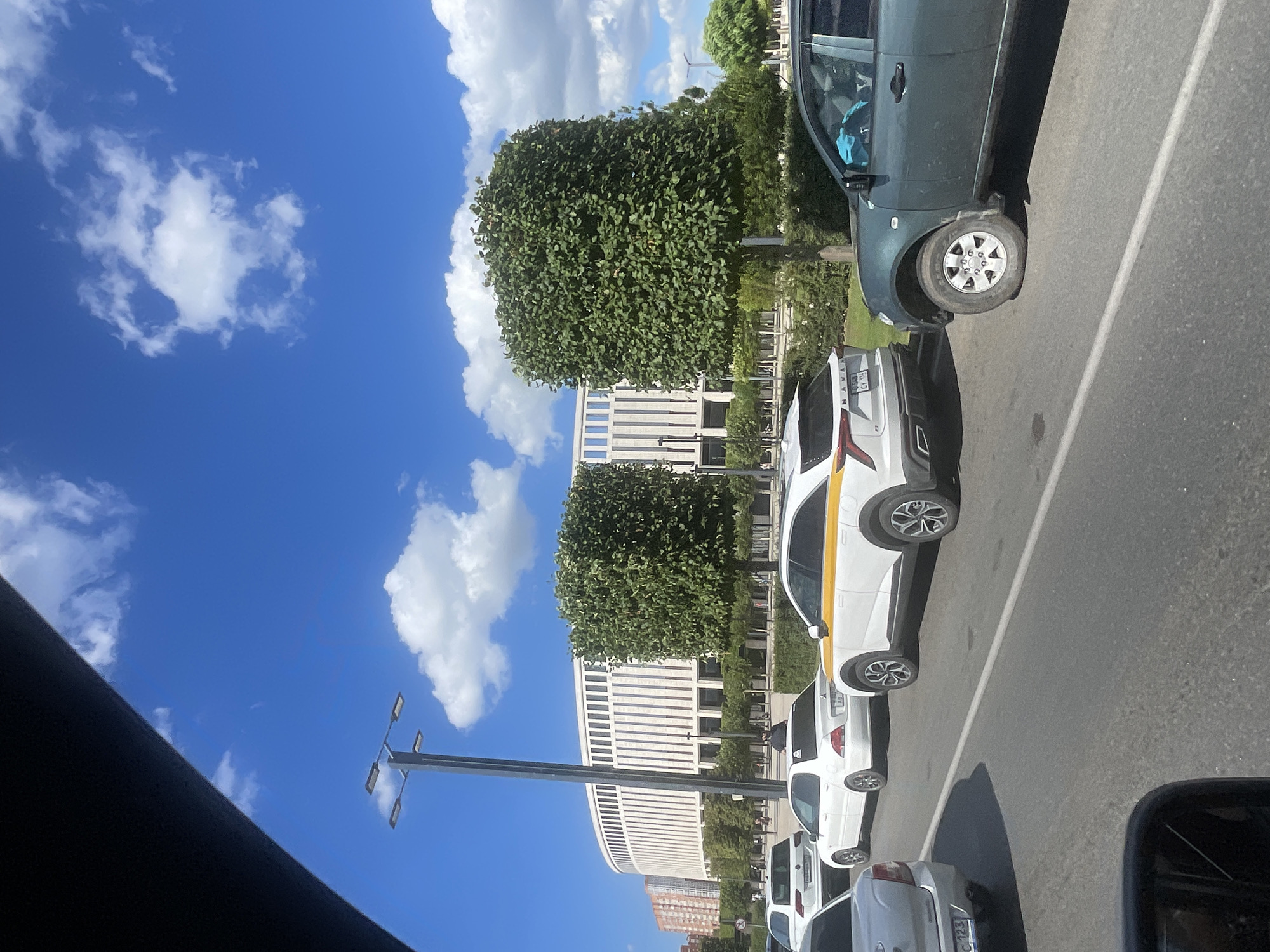 The image shows a bright, sunny day in Krasnodar, Russia, likely near the FKOD arena. In the foreground, several cars are parked or moving along a paved road. A dark green pickup truck is visible on the right, with a blue cloth covering its passenger seat. To its left is a white SUV with a yellow stripe on its roof. Further back, a white sedan and another white car are seen. In the background, a large, modern, cream-colored building with a distinctive curved facade and vertical window panels stands prominently. This building is surrounded by manicured trees and greenery. The sky is a vibrant blue with scattered white clouds. A tall lamppost with multiple lights stands to the left of the road. The overall scene depicts an urban environment with modern architecture and well-maintained public spaces.