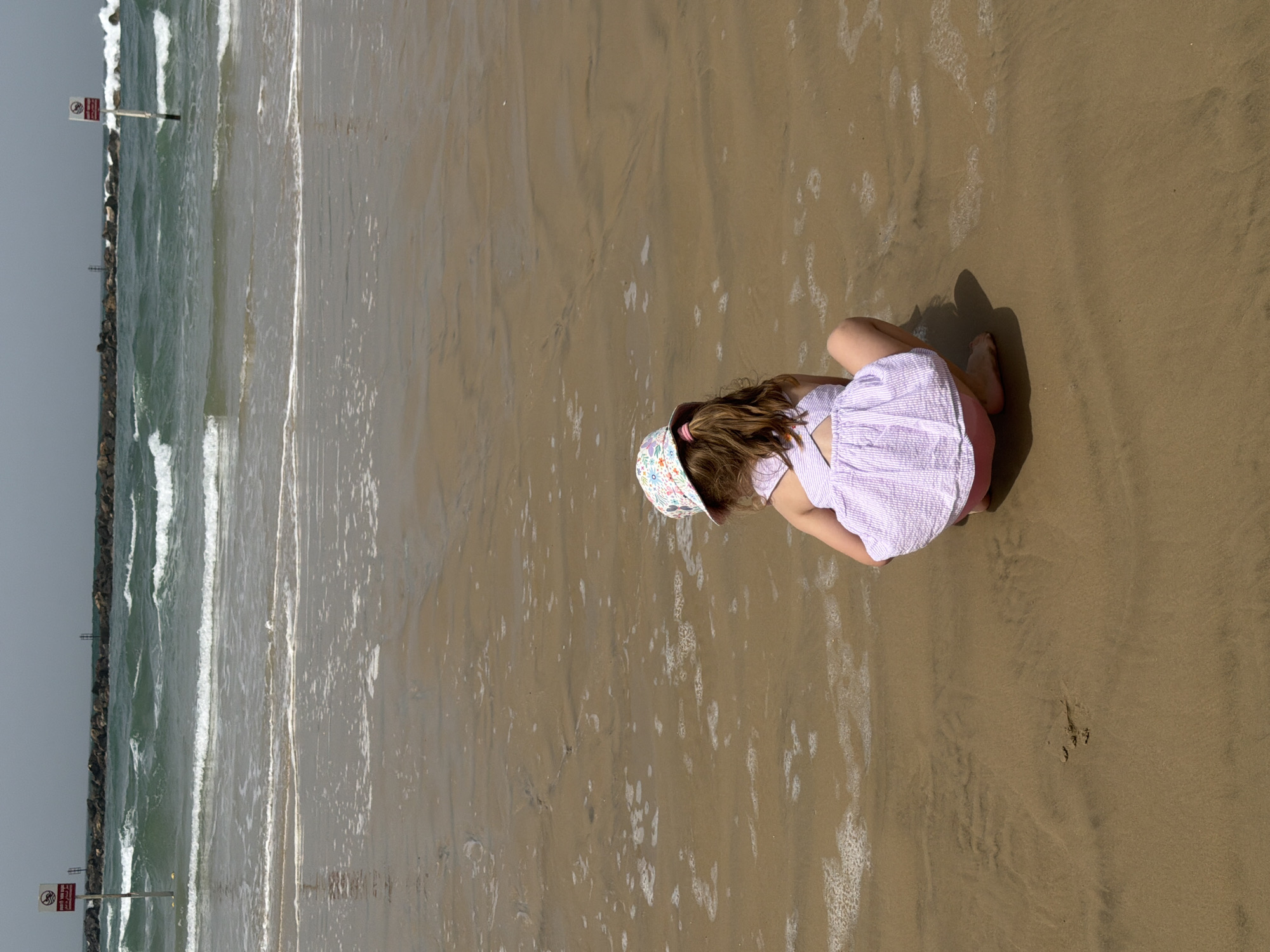 A high-angle, eye-level photograph depicts a child playing on a beach in Tel Aviv, Israel. The child, wearing a light purple and white striped dress and a floral-patterned hat, is kneeling on the wet sand near the shoreline. The beach extends horizontally, with the ocean visible on the left side of the frame. A breakwater constructed of dark rocks and a signpost with a red sign are visible in the ocean near the edge of the frame. The sky is overcast, and the overall lighting is soft and diffused. The sand is wet and reflects the sky, creating a muted, gray-brown color palette. The child appears to be focused on something in the sand, possibly playing or observing.