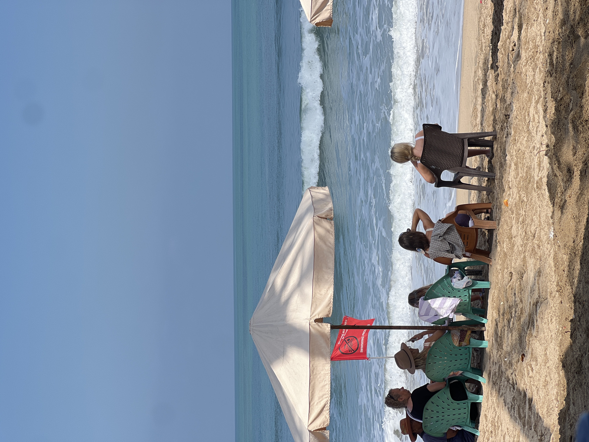 In Kuta, Indonesia, several individuals are seated on beach chairs facing the ocean under a large umbrella. The scene depicts a sandy beach with a calm sea and a clear blue sky. A red flag with a "no swimming" symbol is planted in the sand near the umbrella. The people, all facing away from the camera, appear to be relaxing and enjoying the view. One person is looking at their phone. The overall atmosphere is one of leisure and tranquility.