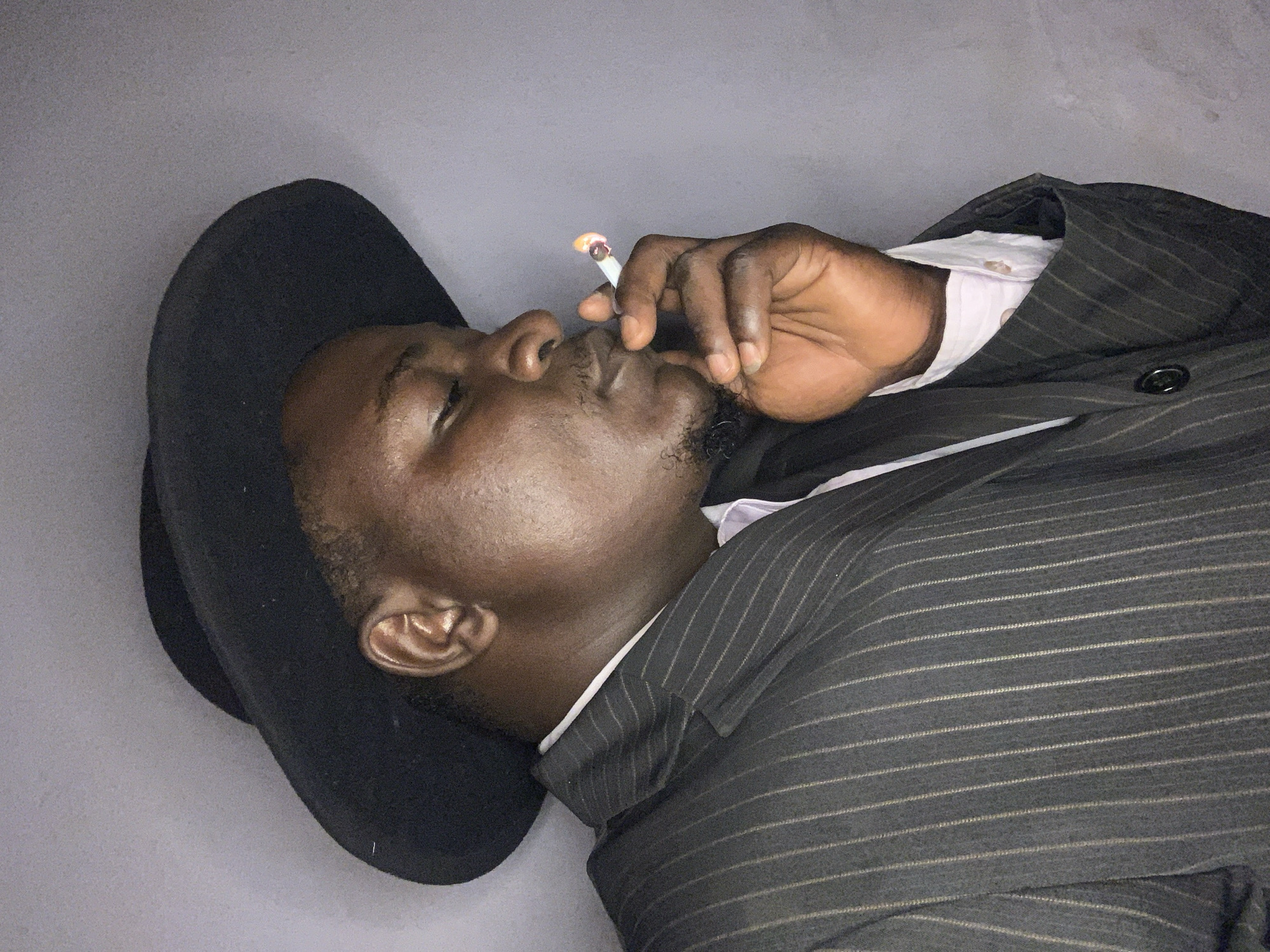 The scene depicts a male individual, appearing to be of Black ethnicity, in the city of Akwanga, Nigeria. He is captured from the chest up in a right-profile view, positioned against a plain, light grey background. The man's eyes are partially closed. He is wearing a black wide-brimmed hat, visible on the top and back of his head, and a dark pinstripe suit jacket over a light-colored collared shirt. His right hand is raised to his mouth, holding a lit cigarette between his thumb and index finger, with the tip exhibiting an orange glow. Facial hair includes a mustache and a short beard on his chin. The lighting appears to be from the front or above.