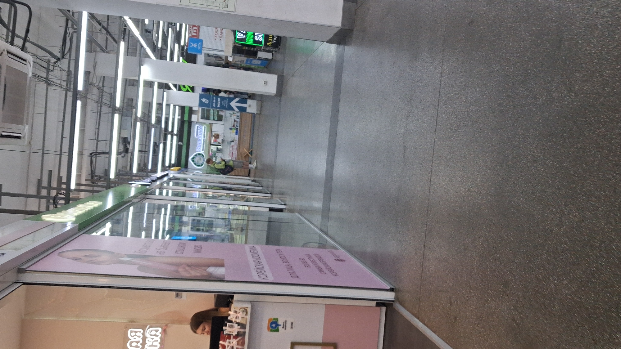 The image shows an indoor shopping mall corridor in Novosibirsk, Russia. On the left side of the image is a retail store with a large glass window displaying products. A woman is visible behind a counter inside the store. The store appears to be selling cosmetics, as indicated by the text in Russian on a large pink sign: "КОРЕЙСКАЯ КОСМЕТИКА" (Korean Cosmetics). The corridor floor is made of polished grey concrete. In the distance, other retail stores and shoppers are visible. The overall environment is brightly lit with fluorescent lighting.
