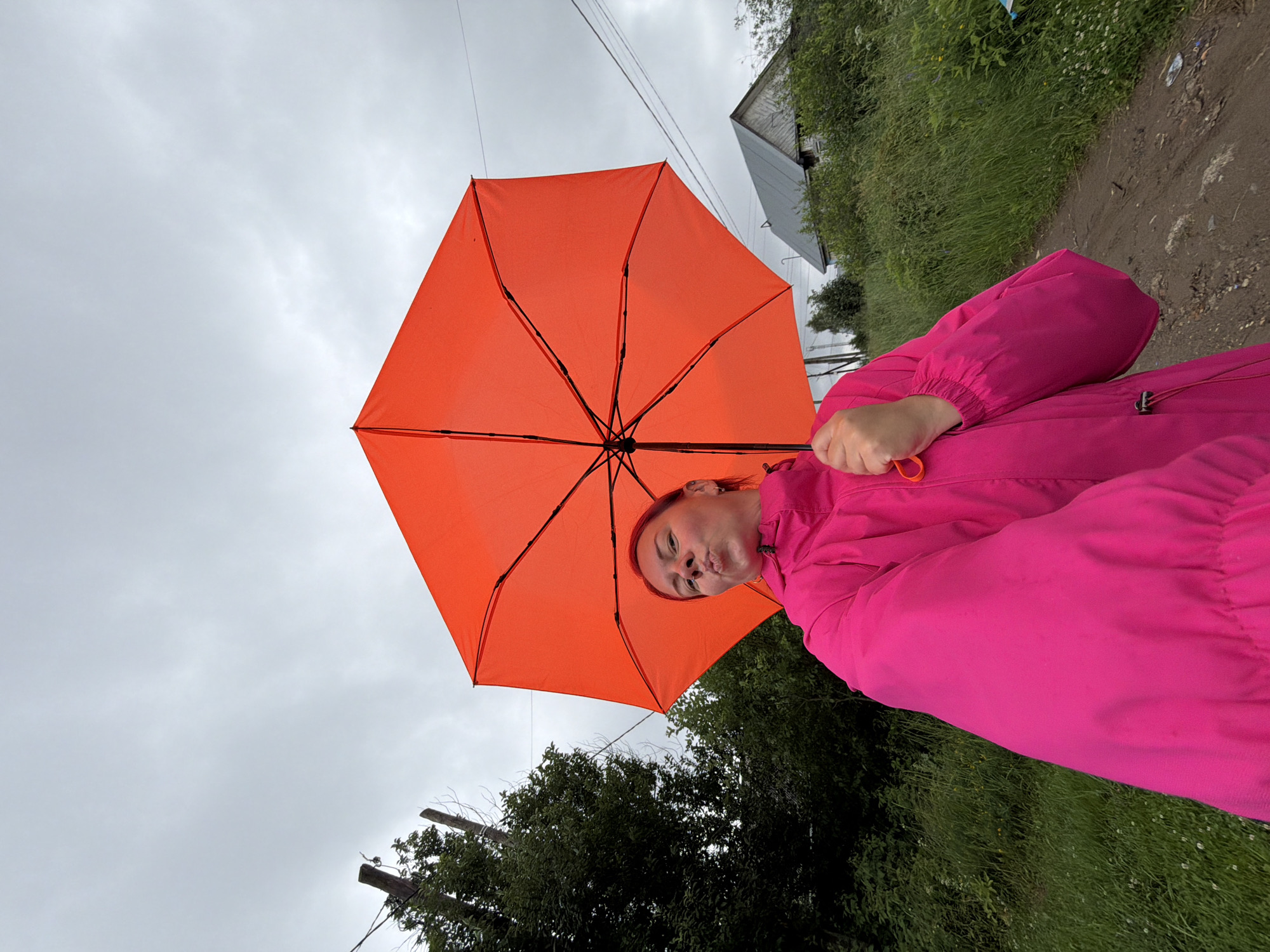A woman with dyed red hair is holding an open, bright orange umbrella and posing for a selfie outdoors. She is wearing a bright pink hooded jacket. The background shows a cloudy sky, green trees, a dirt road, and a weathered wooden house with a gray roof. Power lines are visible across the sky. The scene appears to be in a rural or suburban area, consistent with Chaikovsky, Russia, possibly during overcast weather.
