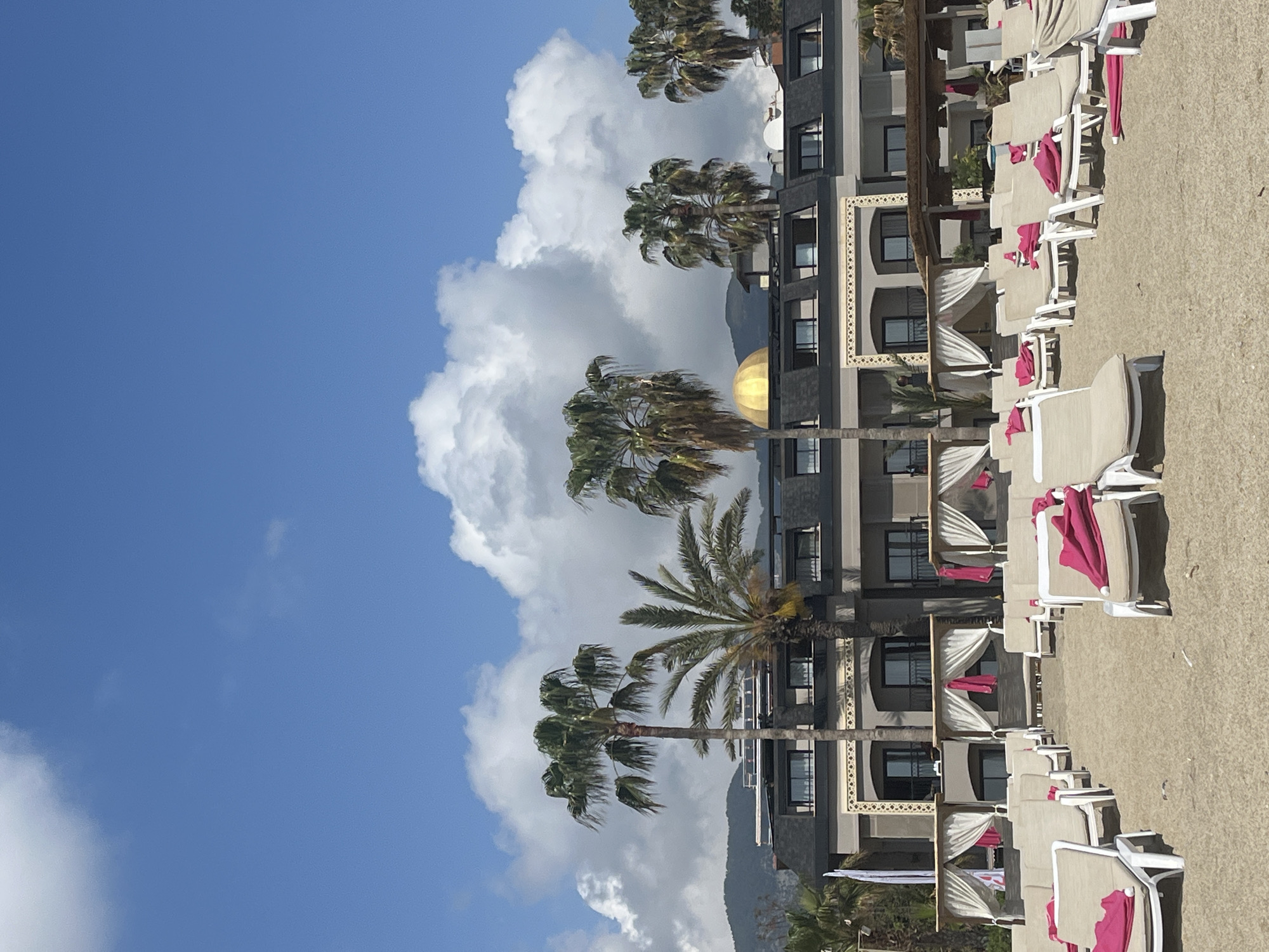 The image captures a vibrant, sunny scene at a resort in Alanya, Turkey. In the foreground, numerous empty sun loungers with light-colored cushions and bright pink towels are neatly arranged in rows on a sandy ground, suggesting a beach or poolside area. Some white-draped cabanas or daybeds are also visible closer to the main building.
The midground is dominated by a multi-story hotel building, characterized by its modern, grey-toned facade with numerous windows and balconies. Several tall palm trees are strategically placed in front of and around the building, their fronds appearing slightly wind-blown. A distinctive golden dome is visible on one section of the building's roof, adding an architectural accent.
In the background, under a bright blue sky, large, fluffy white clouds drift by. Faint outlines of mountains can be seen beyond the resort structures, hinting at the natural landscape of the region.
The scene suggests a peaceful and inviting holiday destination on a clear day, likely morning or early afternoon given the bright lighting. No people are visible, and no specific activities are taking place, though the setup of the loungers and cabanas clearly indicates leisure and relaxation. There is no visible text in the image.